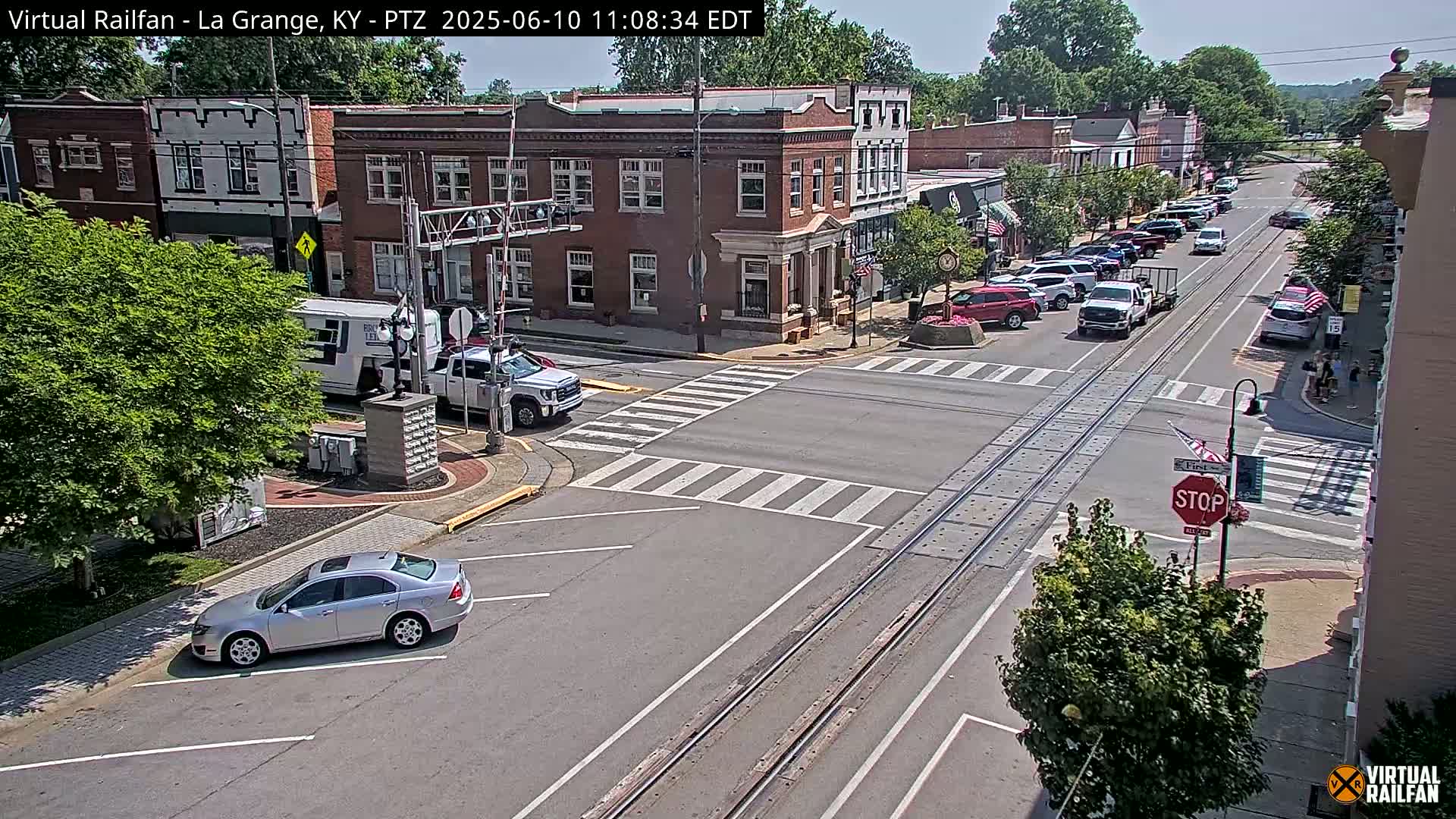 A sunny day in a small town features a street intersection with parked cars, a few moving vehicles, and brick buildings, all situated near a set of train tracks.
