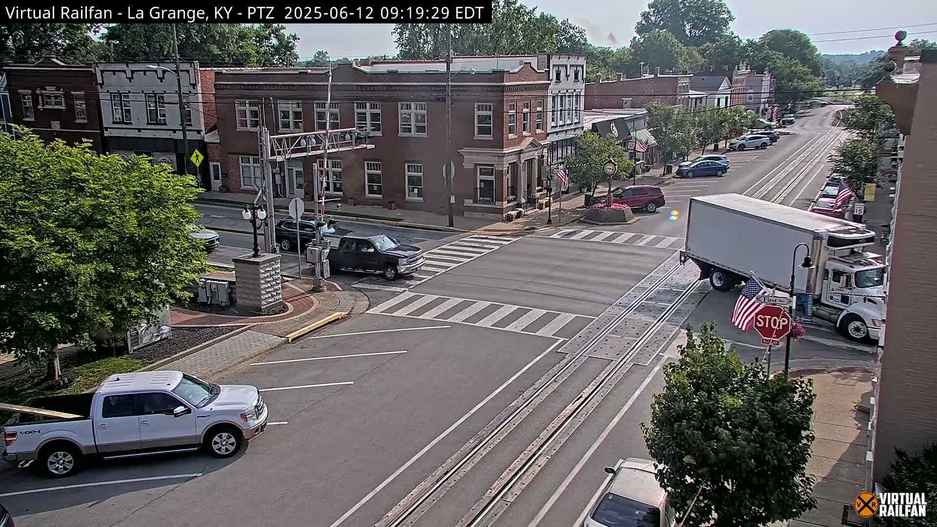 A mostly sunny day shows a small town intersection with several vehicles, including a pickup truck and a large semi-truck, at a railroad crossing.
