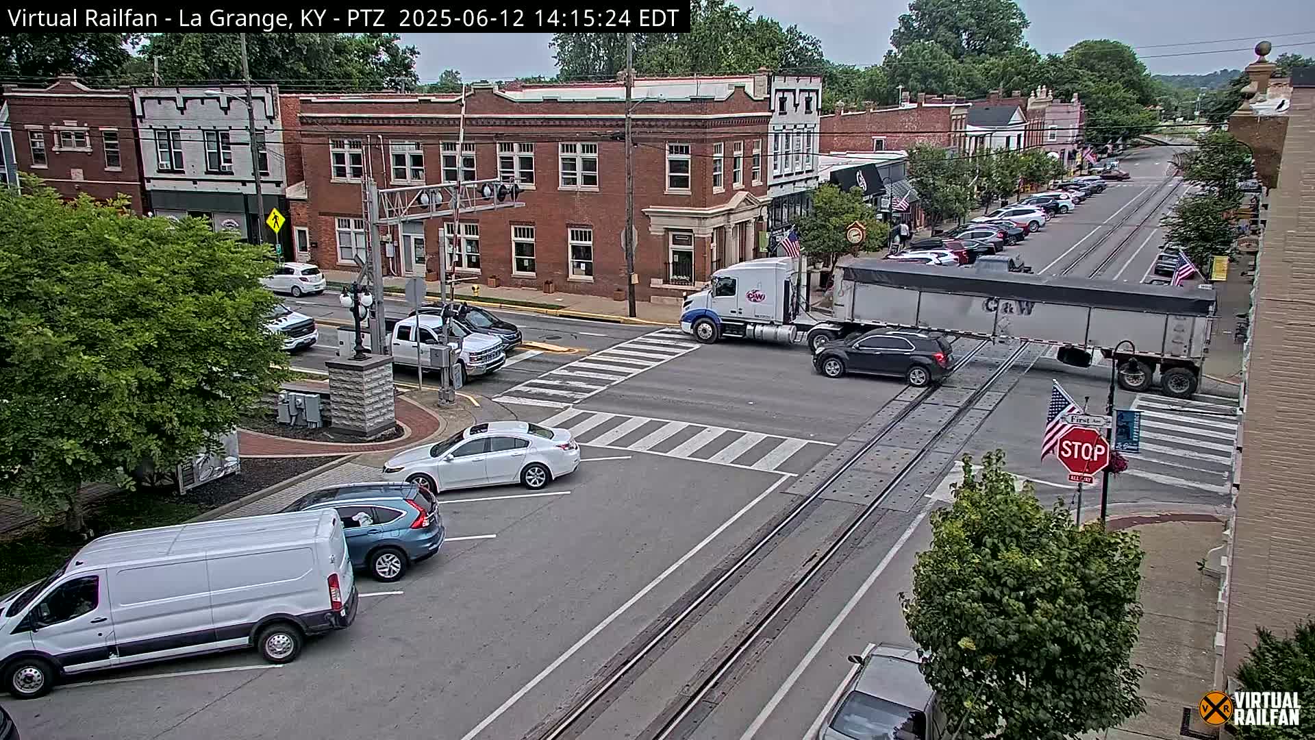 A partly sunny day in a town square shows several vehicles at a crosswalk and intersection with railroad tracks; a large semi-truck is stopped at the tracks.