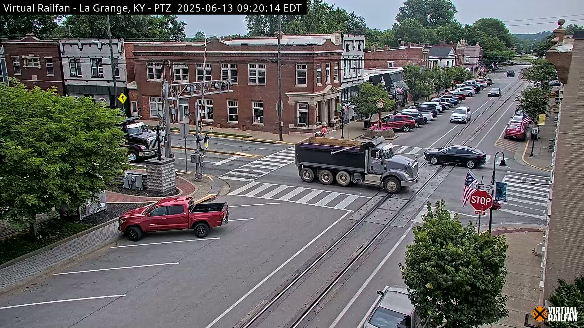 A dump truck and several other vehicles are at an intersection in a town on a partly cloudy day.