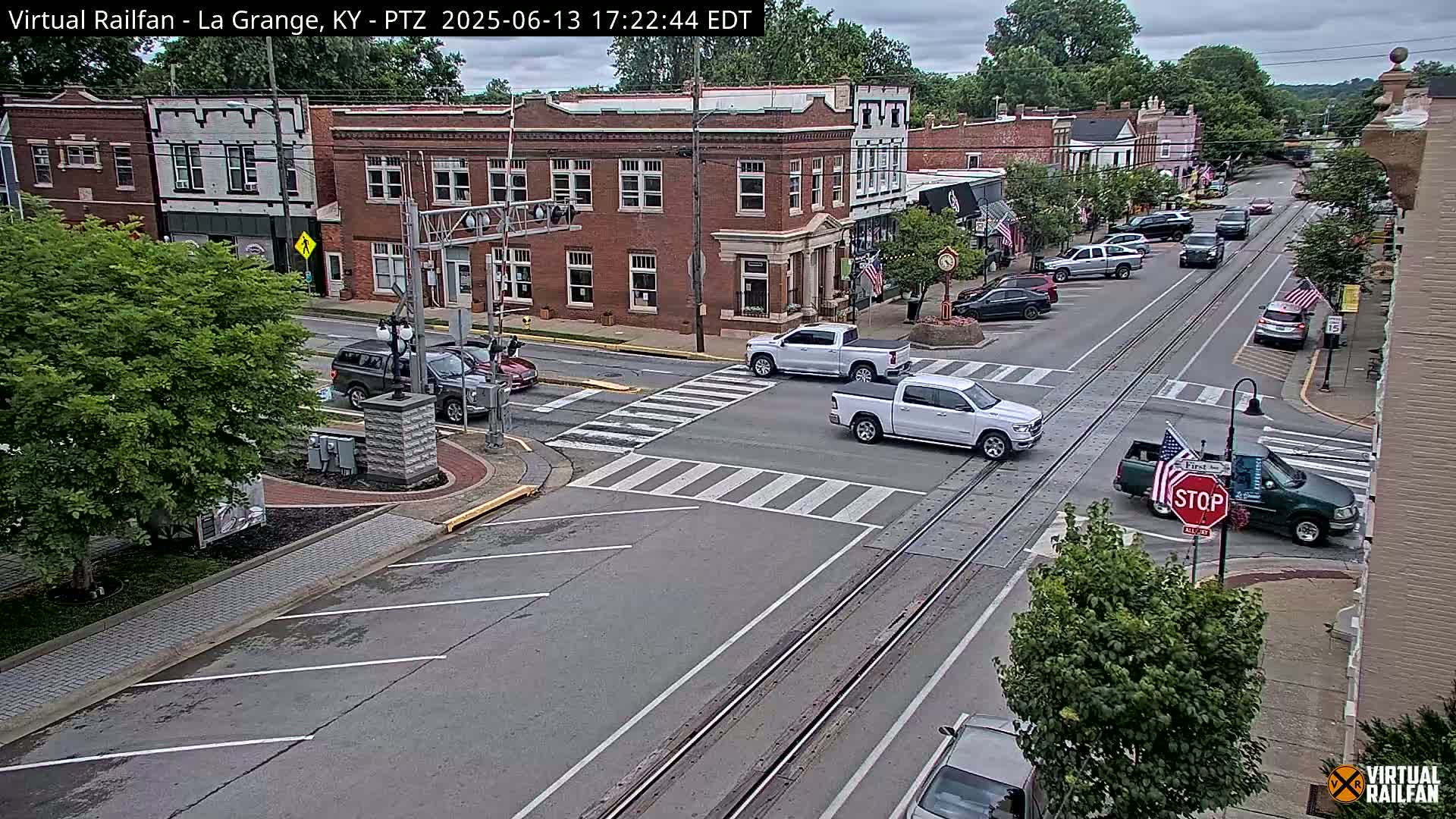 A mostly sunny view of a town's street intersection with several vehicles, including trucks and cars, and brick buildings lining the street.