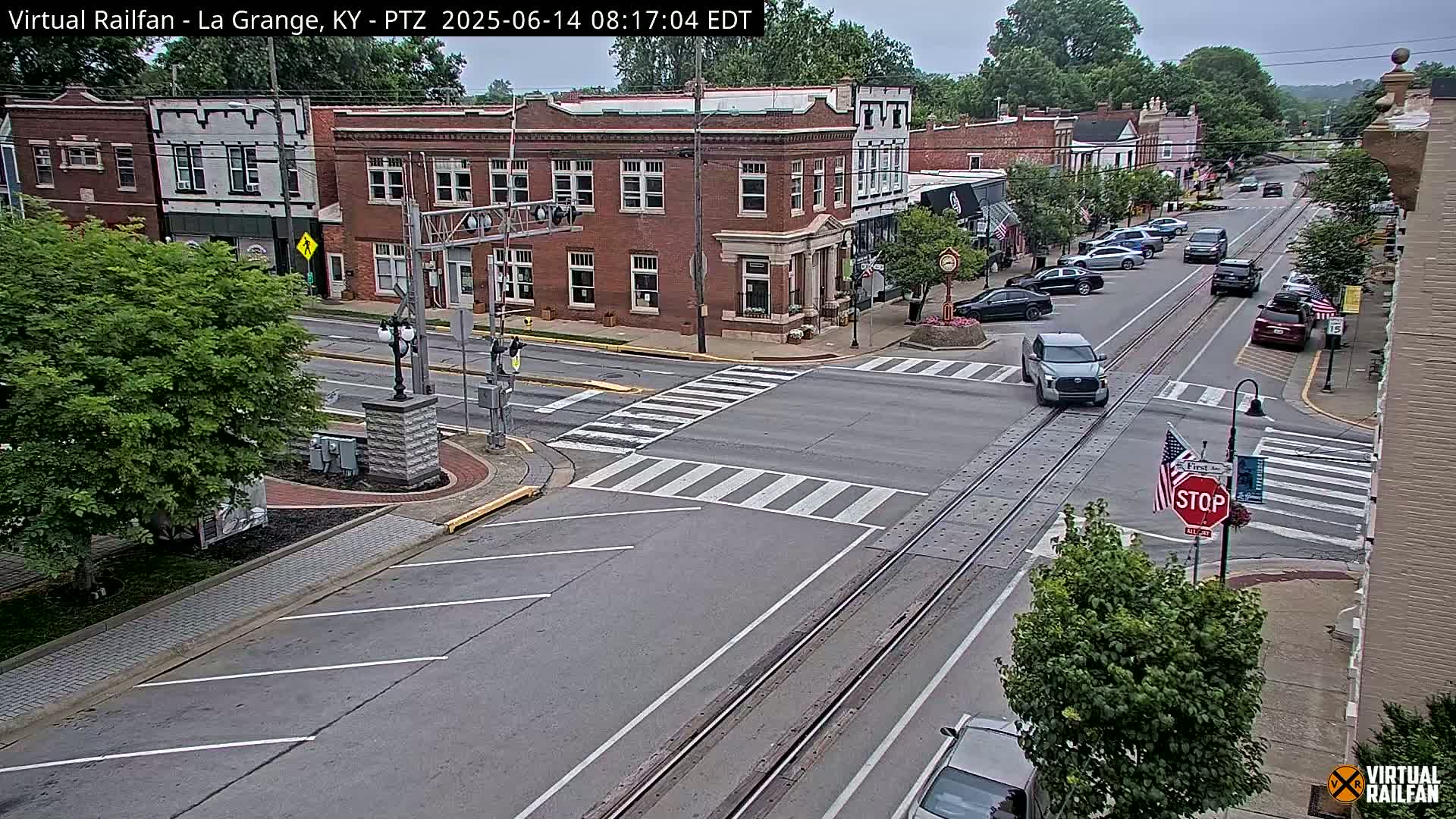 A mostly cloudy day shows a view of a town's street intersection with brick buildings, cars, and train tracks running down the center of the road.