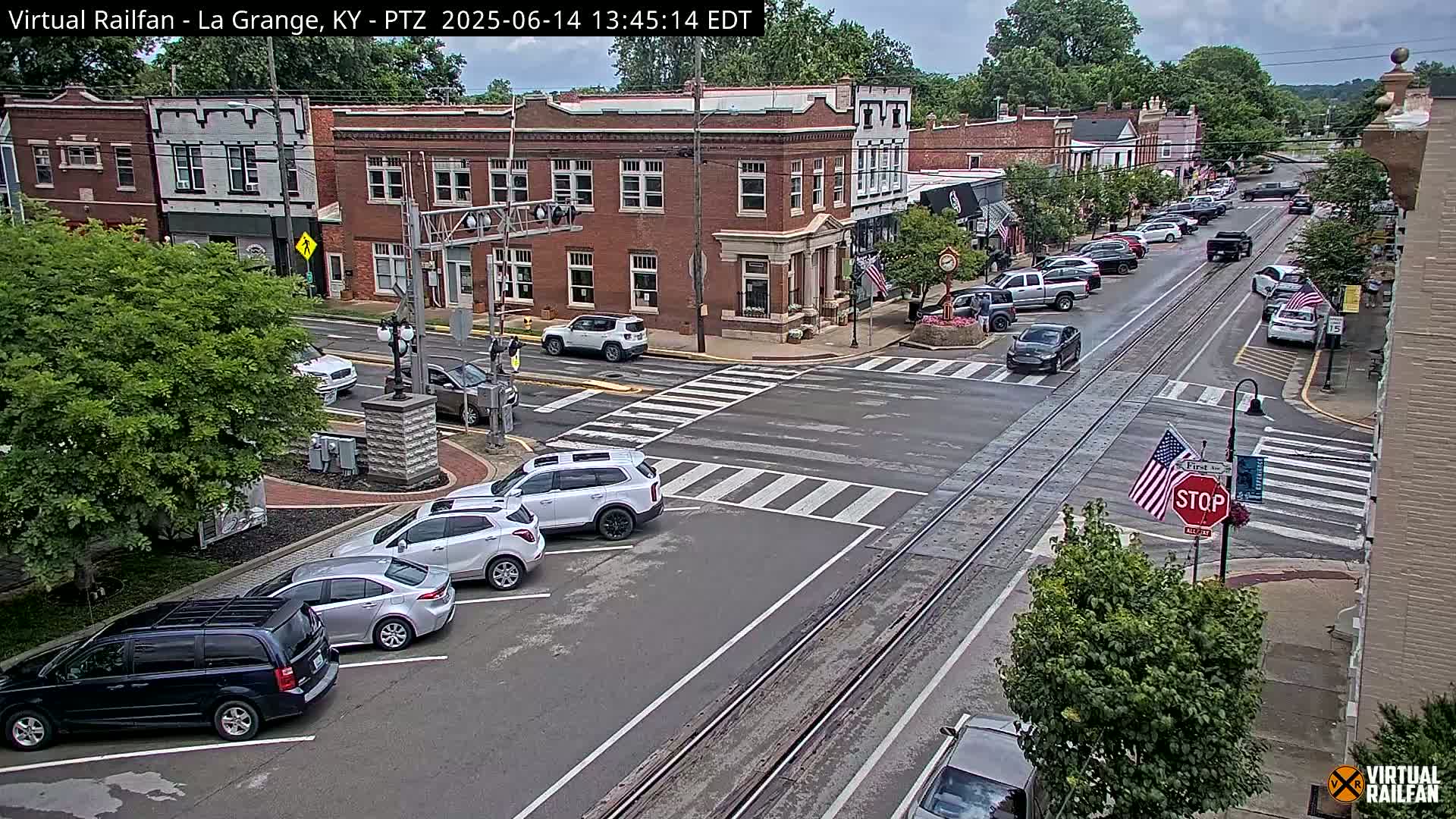 A partly cloudy day shows a street scene in a town with several brick buildings, parked cars, and a few cars driving on a road with train tracks running through it.