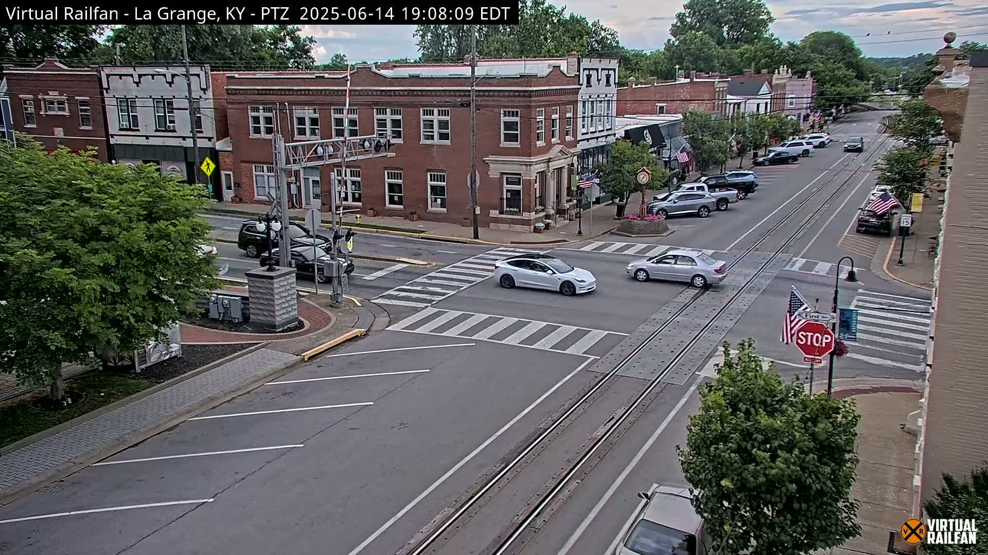 A partly cloudy day shows a street scene in a town with several brick buildings, parked cars, and a few cars driving on a road with train tracks running through it.