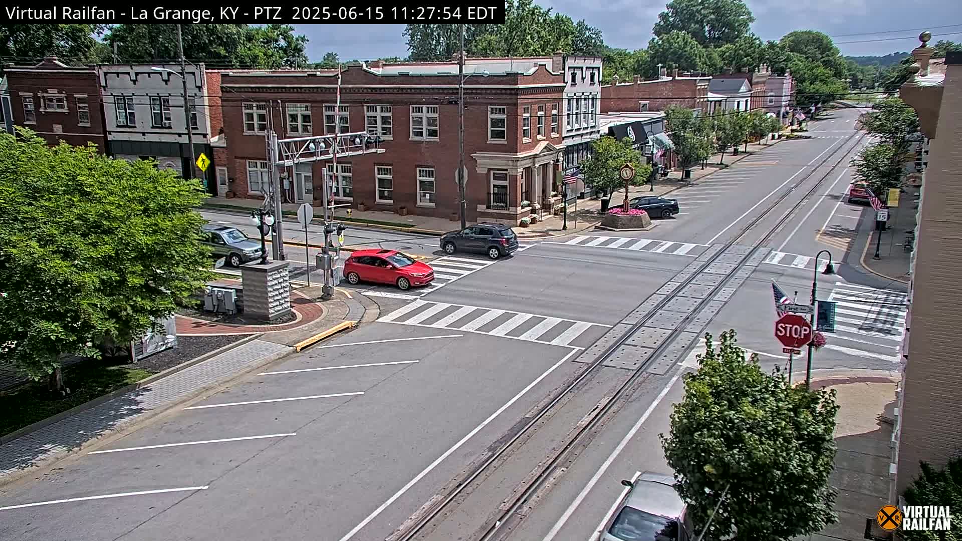 A mostly sunny day reveals a town's street intersection with brick buildings, a few cars, and train tracks running down the road's center.