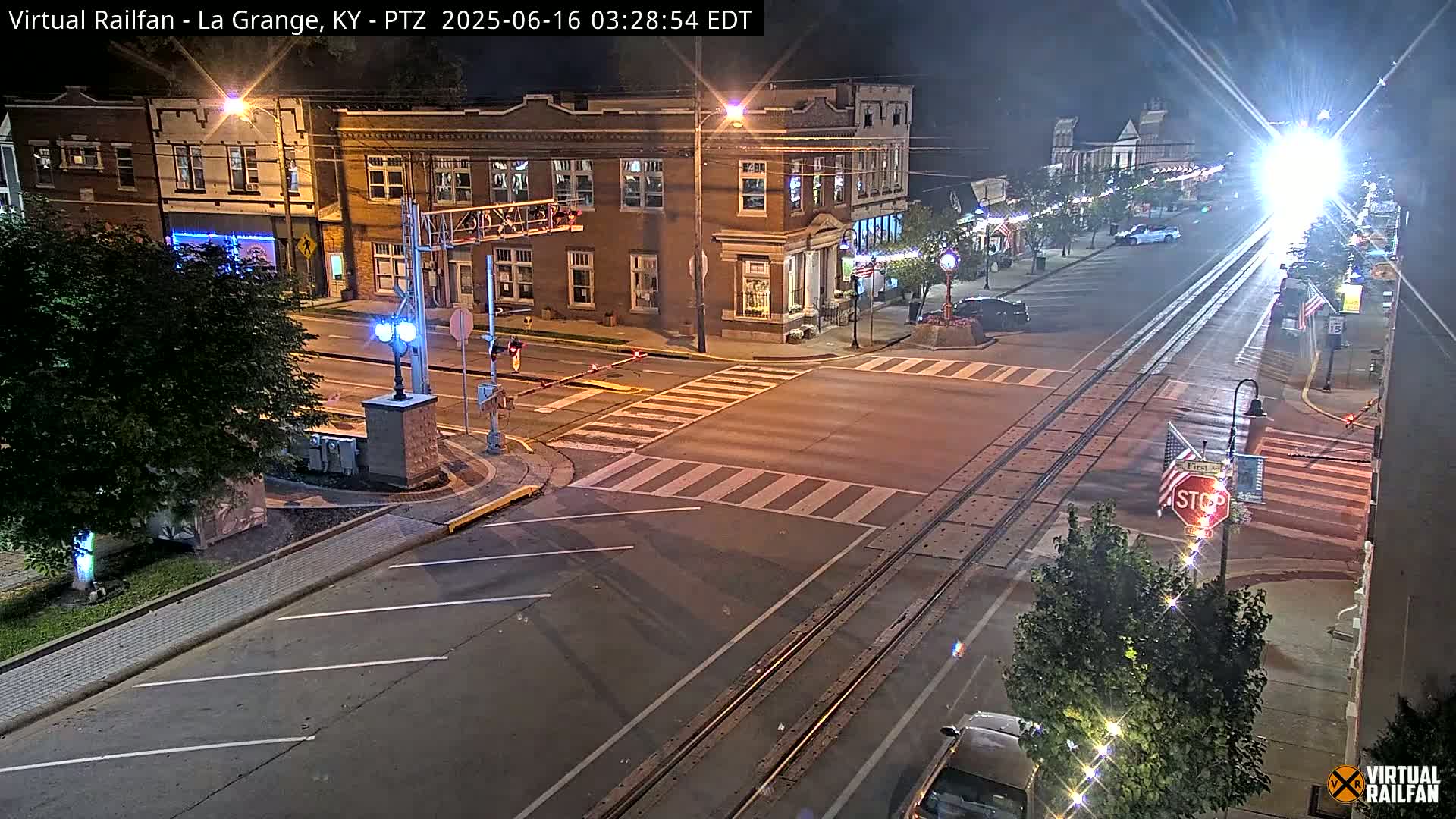 A nighttime view of a street intersection with train tracks, brick buildings, and a few cars under clear skies.