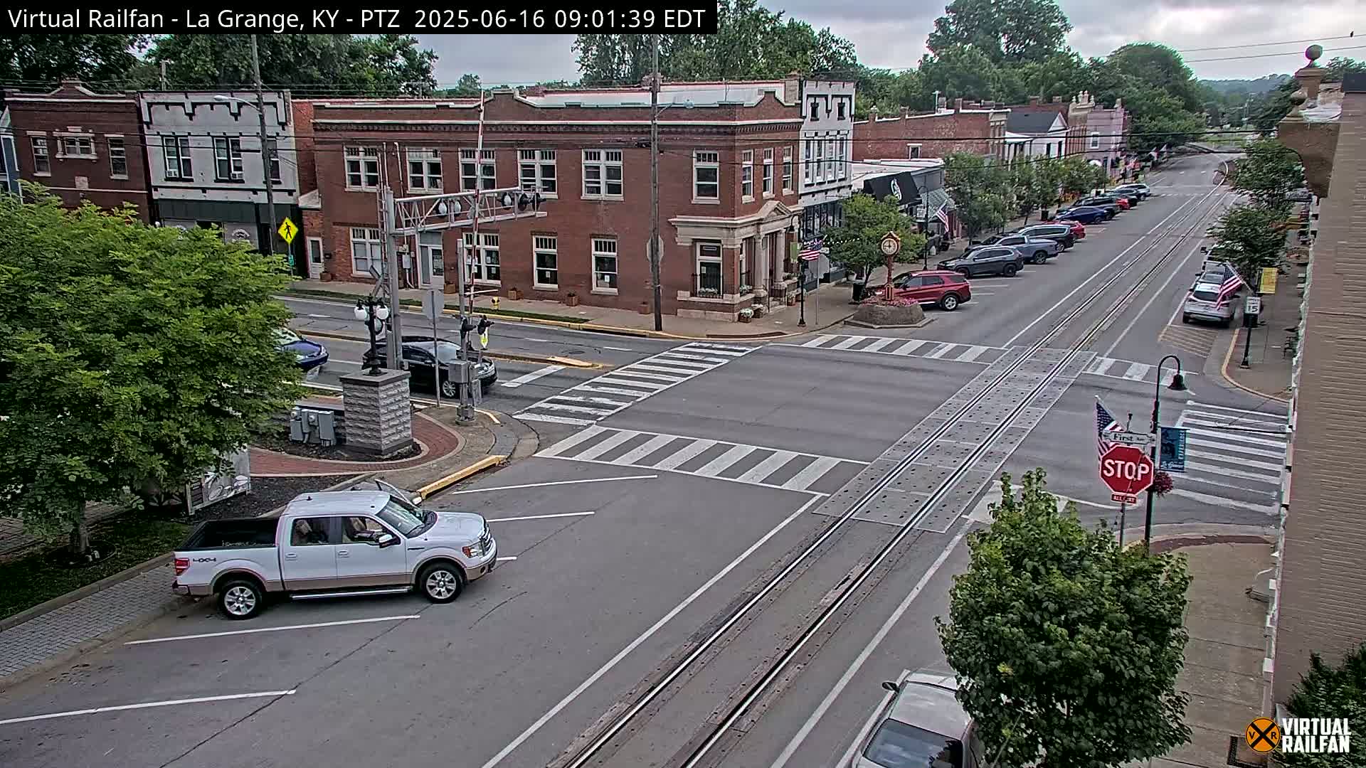 A partly cloudy day shows a street scene in a small town with parked cars lining a street that intersects with another street at a crosswalk, and a white pickup truck parked in a nearby lot.