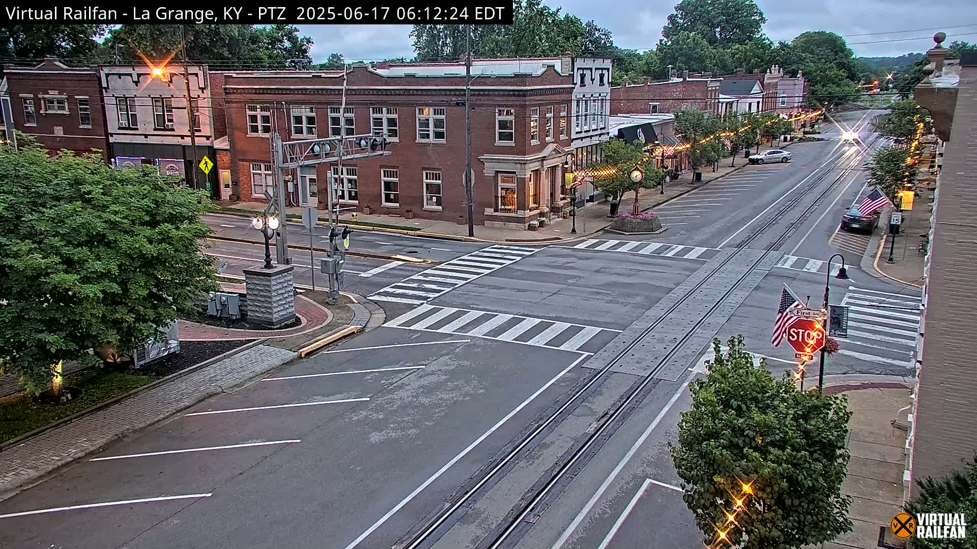 A mostly overcast view of a town's street intersection with brick buildings and parked cars, featuring train tracks running down the center of the road.