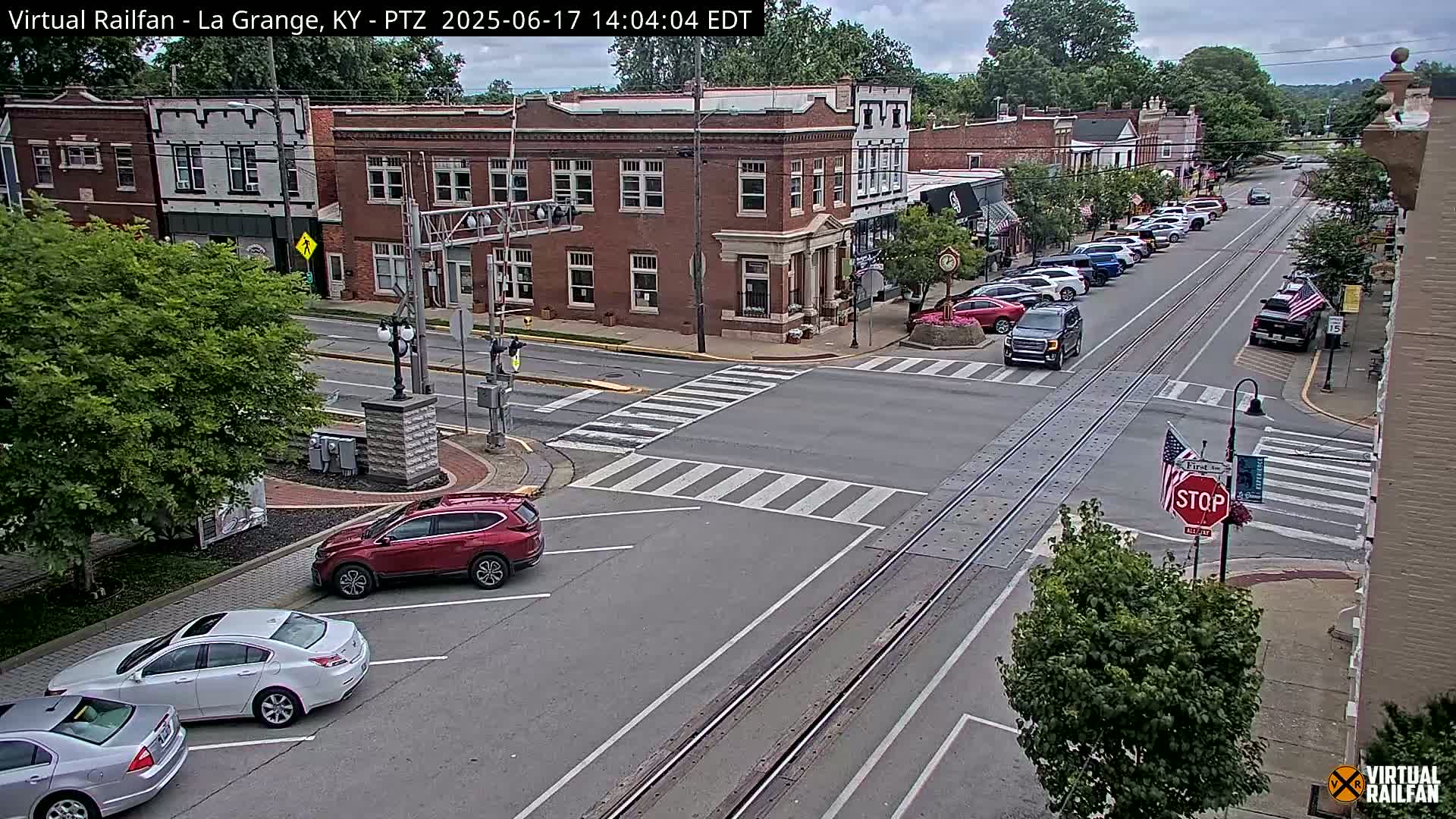 A mostly sunny day in a small town features a street scene with parked cars, several cars at an intersection, and railroad tracks running down the center of the street.