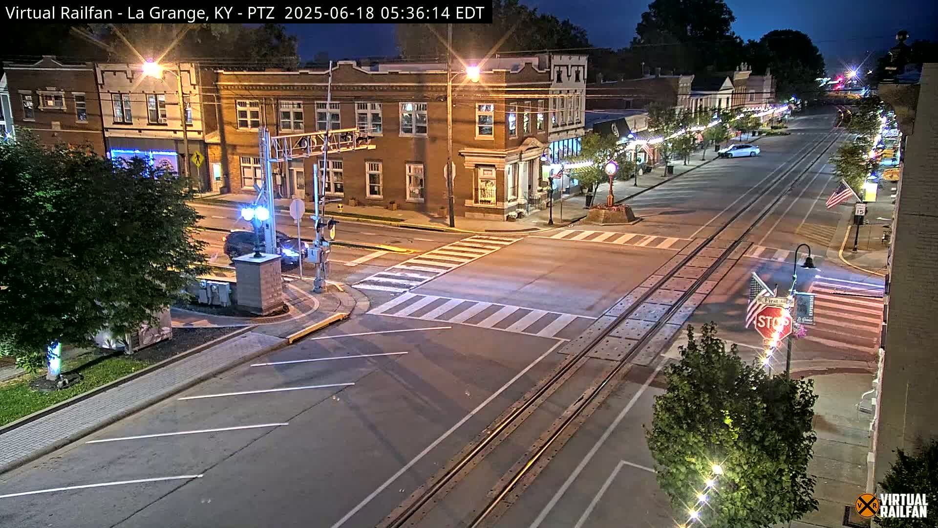 A nighttime street scene shows a brick building at the intersection of a road with train tracks, under a mostly clear sky.