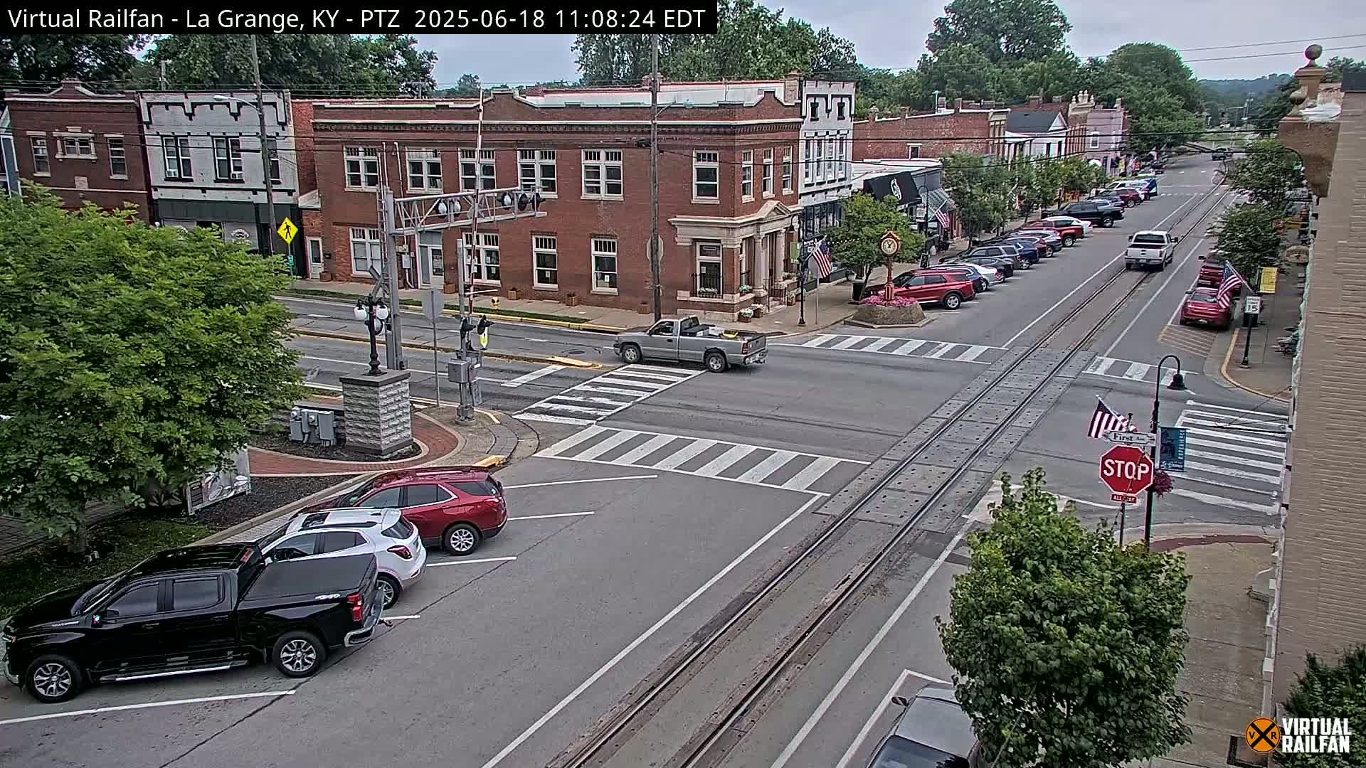 A partly cloudy day reveals a town's main street intersection with parked cars, a few moving vehicles, and brick buildings.