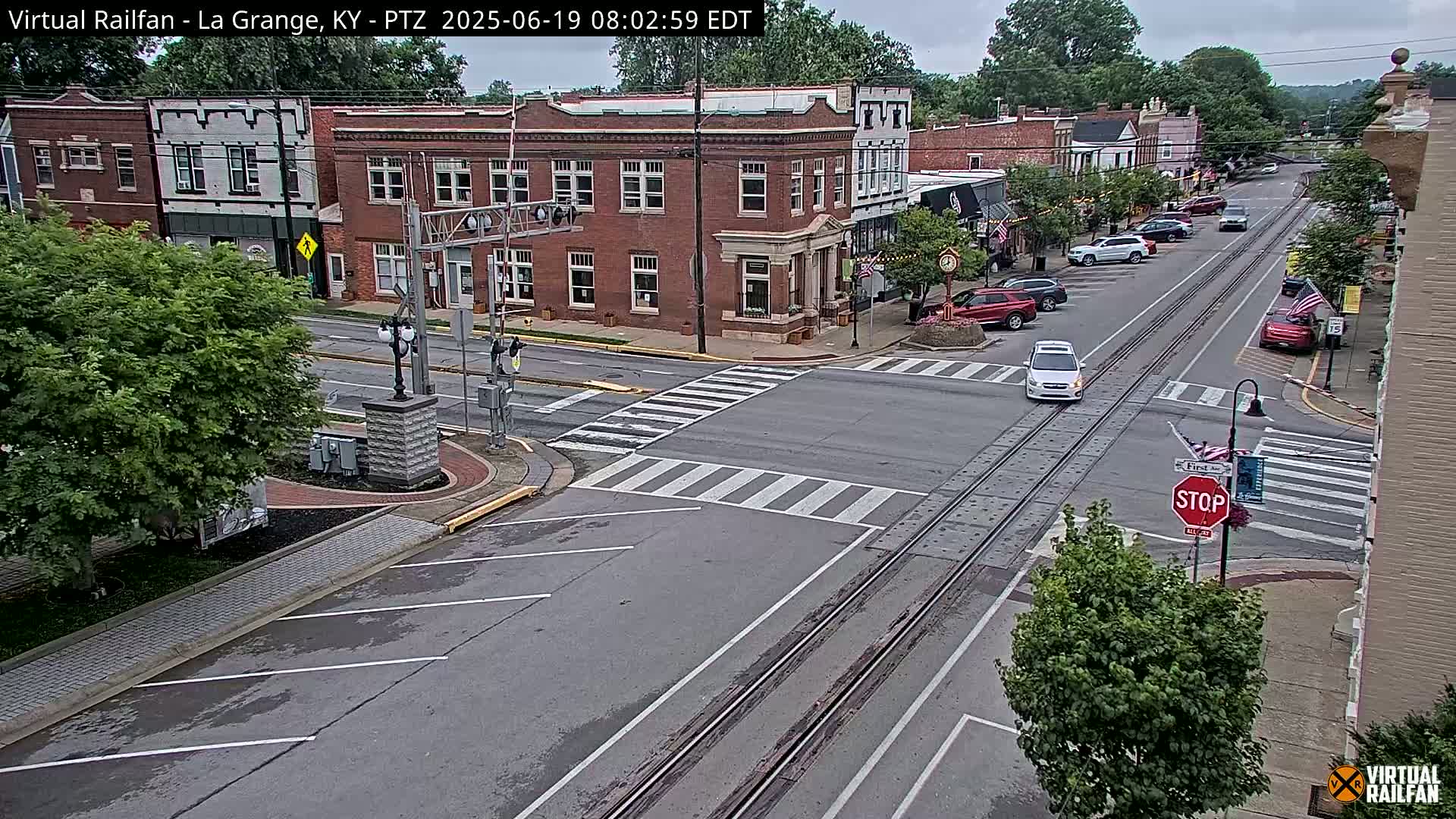 A partly cloudy day shows a view of a street intersection in a small town, with brick buildings lining the street and several cars parked along the curb.