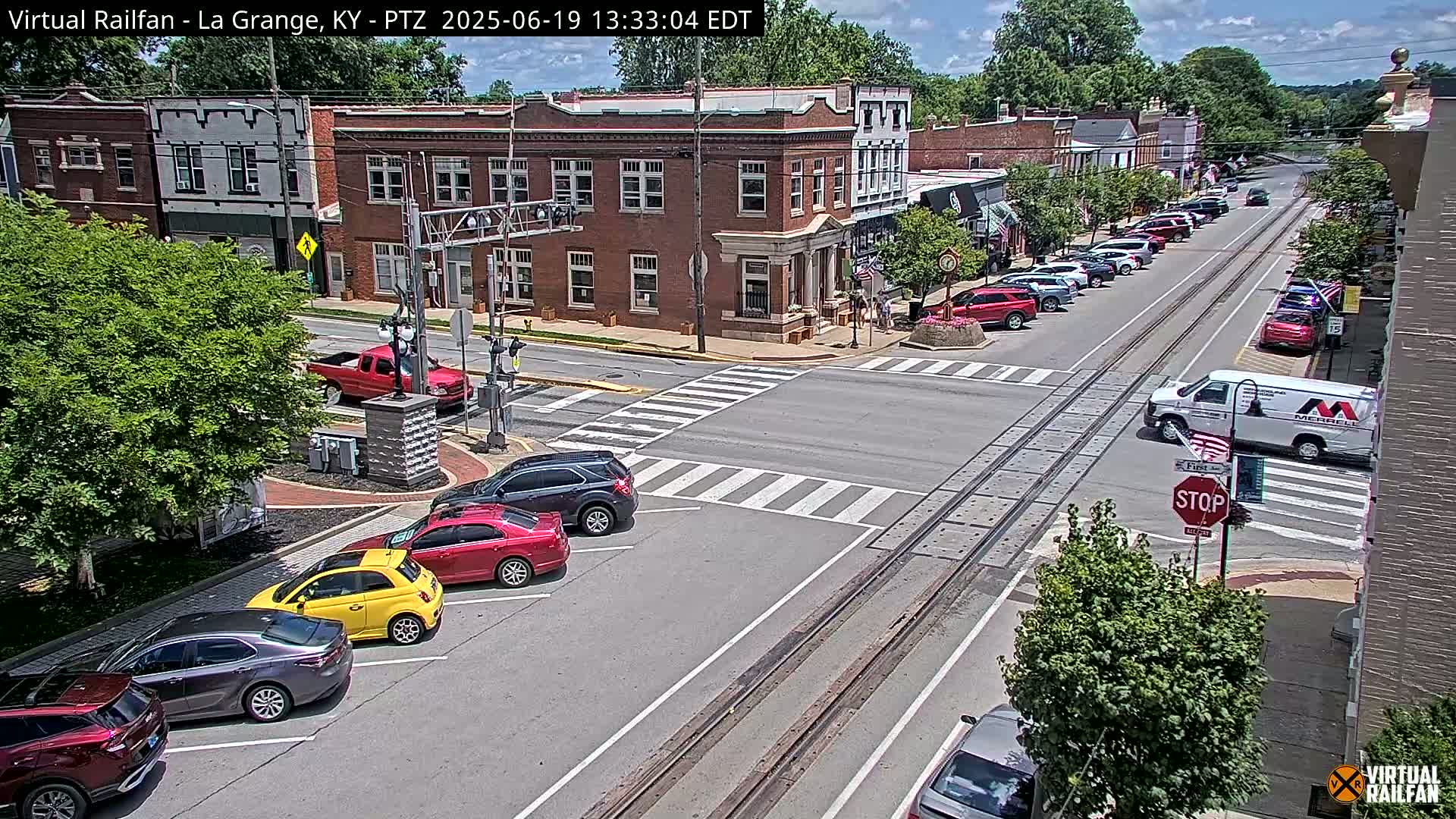 A sunny day reveals a town square with parked cars lining the street, several cars at a crosswalk, and a white van near a stop sign in front of brick buildings.