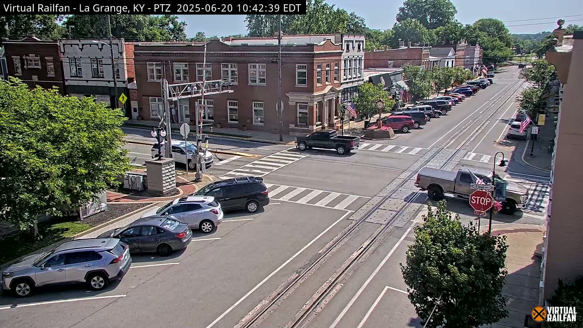 Cars are parked along the street next to brick buildings on a sunny day, with a railroad track running down the center of the road.