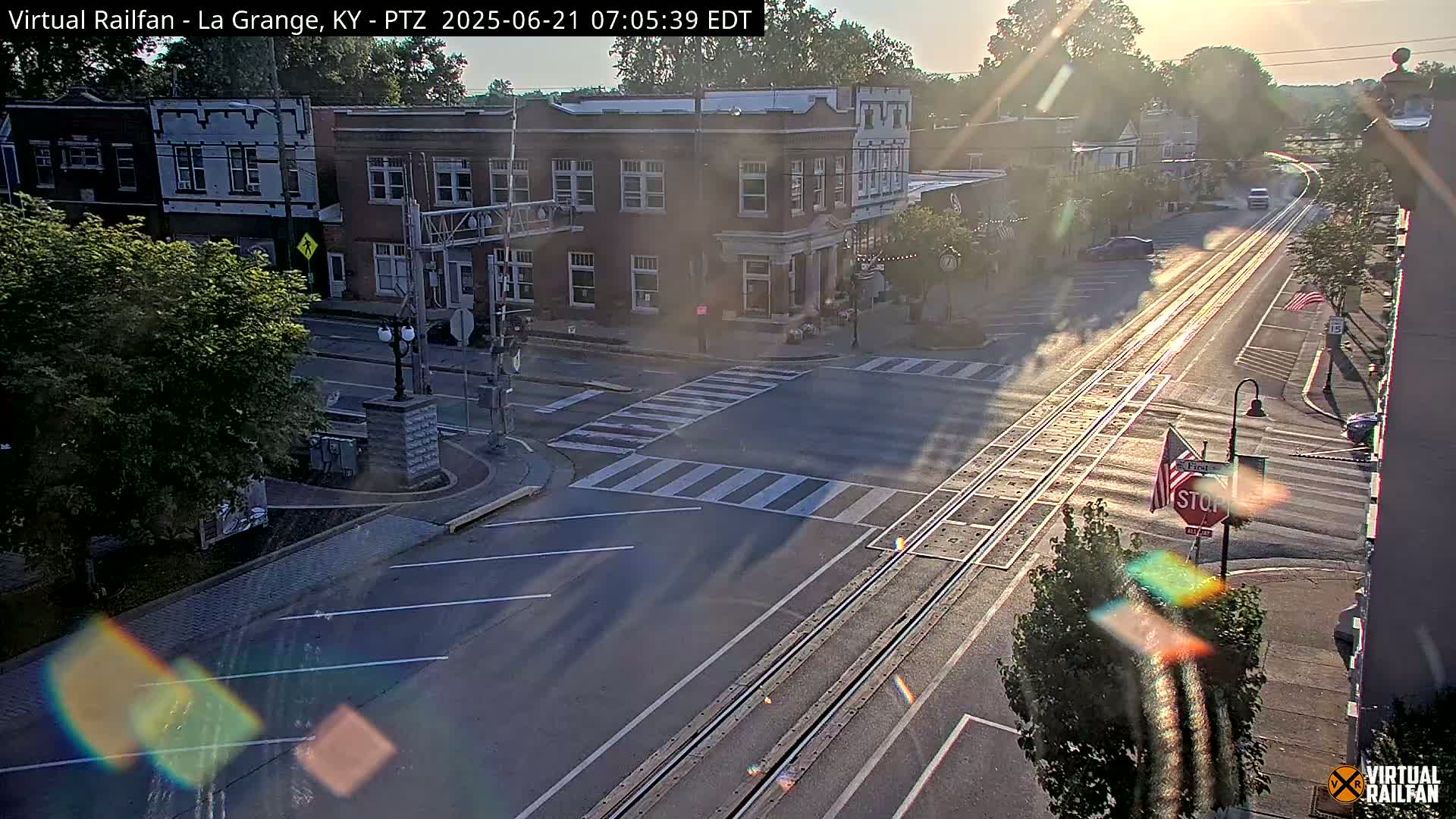 A sunny morning scene shows a street intersection in a small town with train tracks running down the middle of the road, brick buildings lining the street, and a few cars parked along the sides.