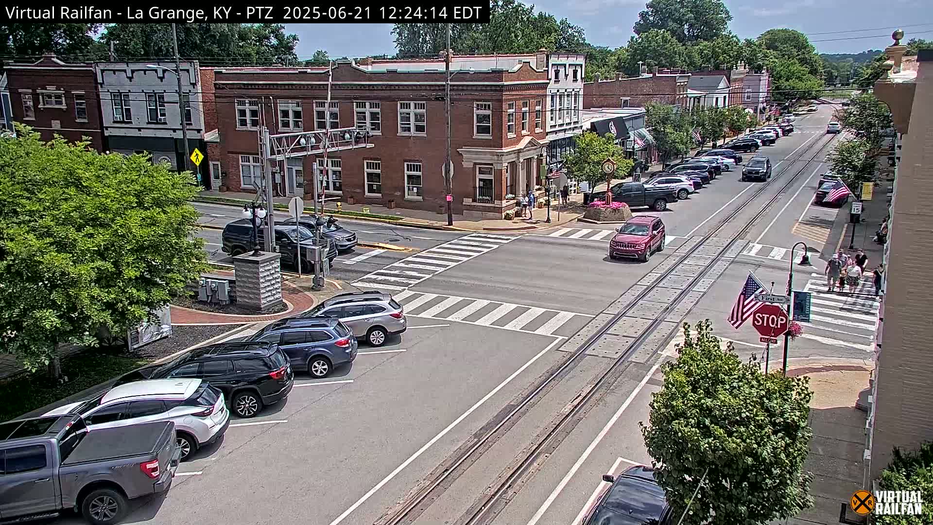 A sunny day in a town with brick buildings, parked cars lining the street, and a few vehicles at a crosswalk.