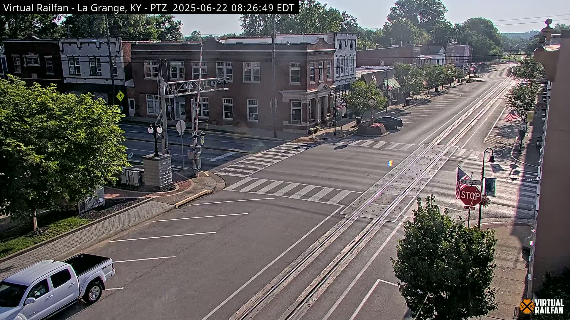 A mostly empty street with railroad tracks running down the center, flanked by buildings and trees under sunny skies.