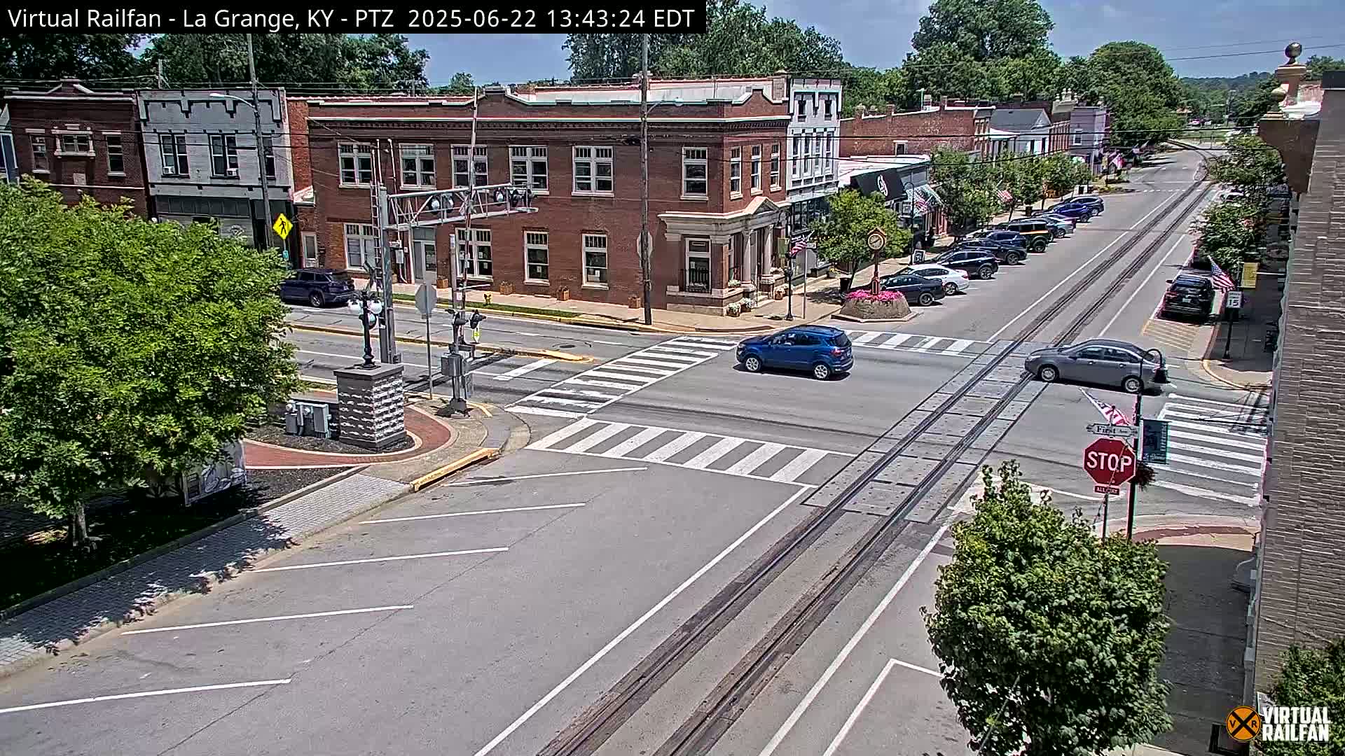 A sunny day in a small town with several cars at an intersection, brick buildings lining the street, and train tracks running down the middle of the road.