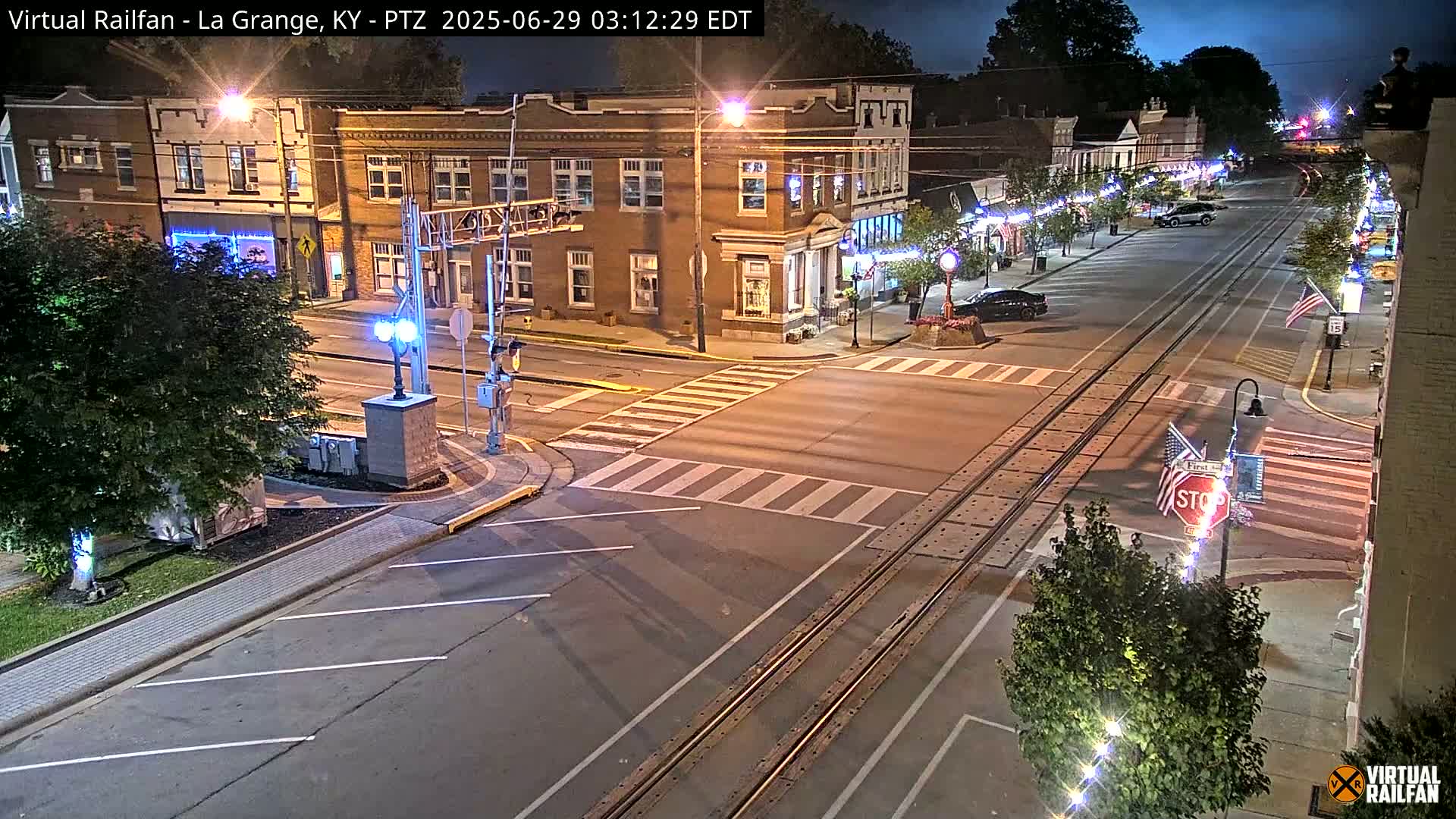 A nighttime street scene shows a town square with a railway track running down the center of the road, intersected by crosswalks and streetlights.