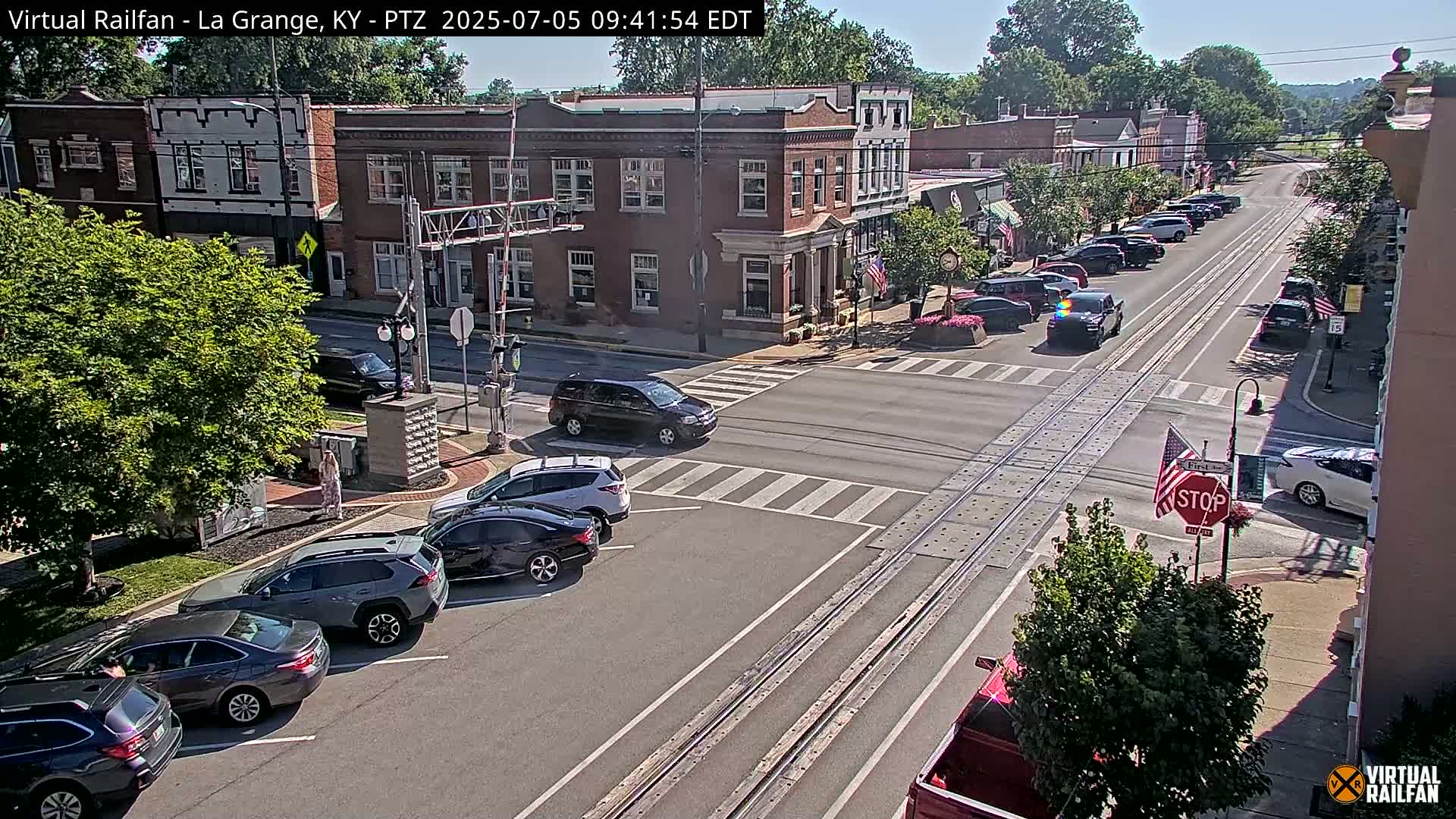 A sunny street scene shows a town square with several parked cars and a few cars driving, alongside brick buildings and lush green trees.