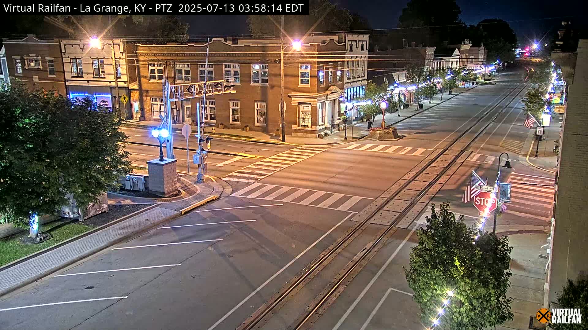 A nighttime street scene shows a quiet intersection with brick buildings, streetlights, and train tracks running down the center of the road.