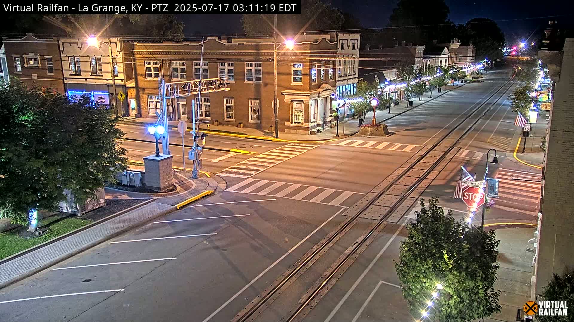 A nighttime view of a town's street intersection with train tracks, showing brick buildings, streetlights, and crosswalks.