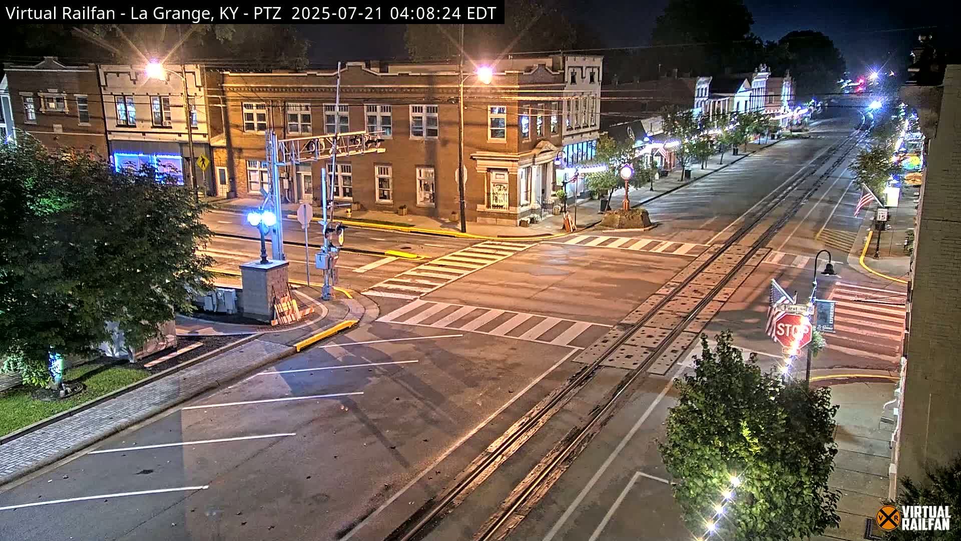 A nighttime view of a street intersection in a town, with brick buildings lining the street, train tracks crossing the road, and a stop sign visible.