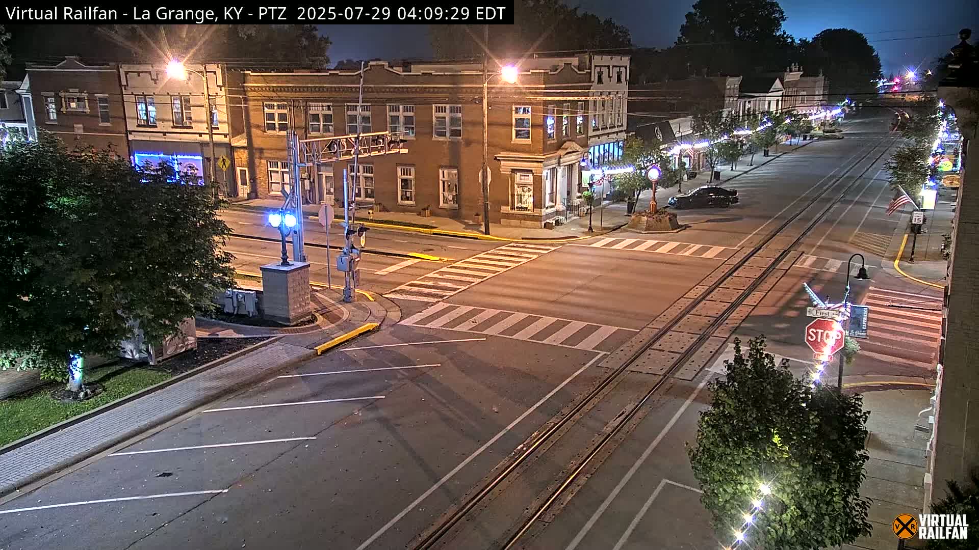 A nighttime street scene shows a quiet intersection with train tracks, brick buildings, and sparsely parked cars under a clear sky.
