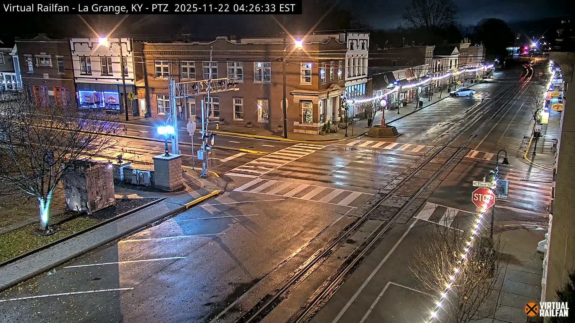 A rainy night scene shows a small town street lined with historic buildings, bare trees adorned with string lights, and prominent railroad tracks running through a reflective, wet intersection where a single white car is parked.