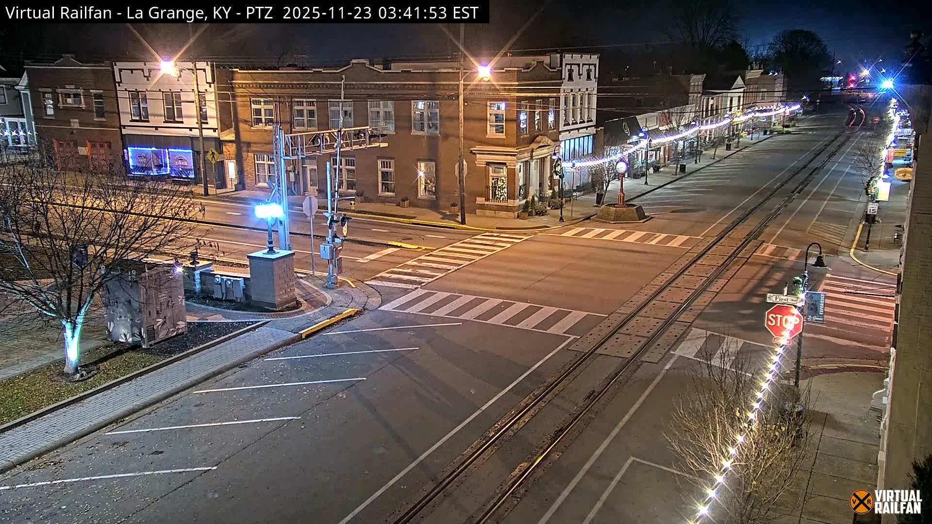 A quiet downtown street at night, illuminated by numerous streetlights and festive string lights adorning buildings and trees, features a prominent railroad track running down its center, all under clear skies.