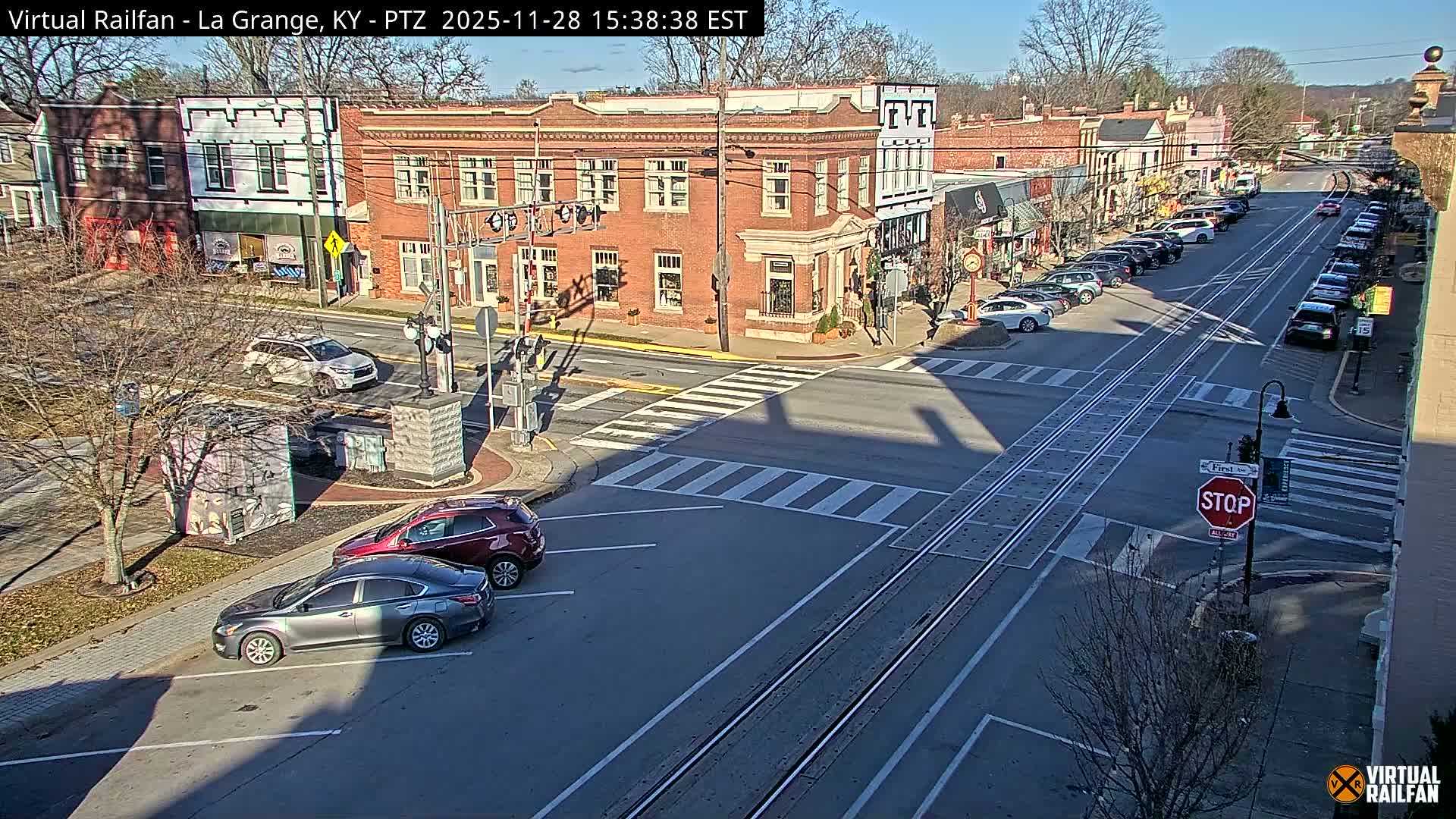 On a clear, sunny day, a bustling town street features numerous parked cars, several brick buildings lining the roads, and prominent railroad tracks running down its center, with bare trees visible under a bright blue sky.