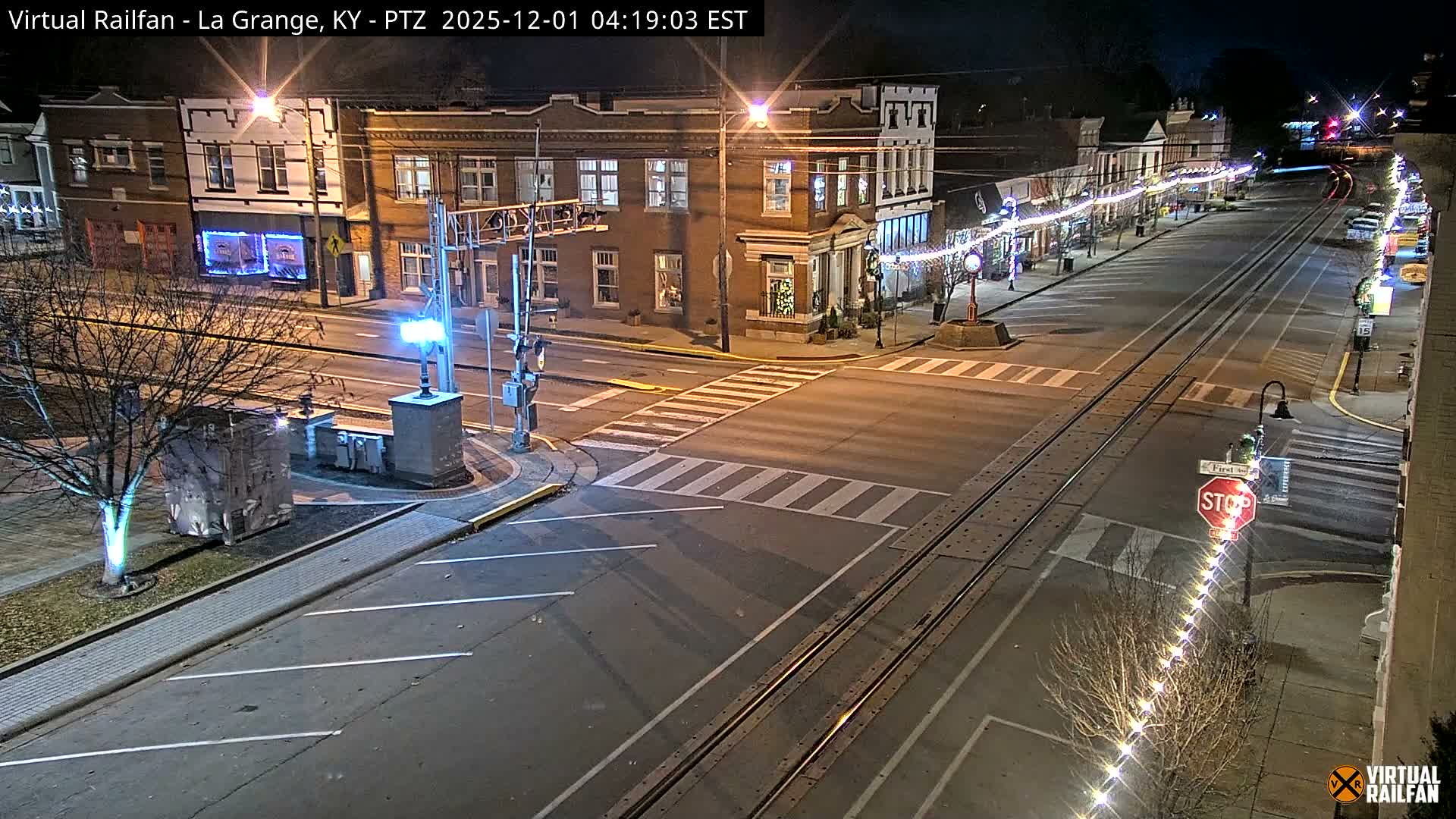 An overhead view on a clear, dark night captures a quiet downtown street with train tracks running down its center, flanked by historic brick buildings decorated with string lights and bare trees.