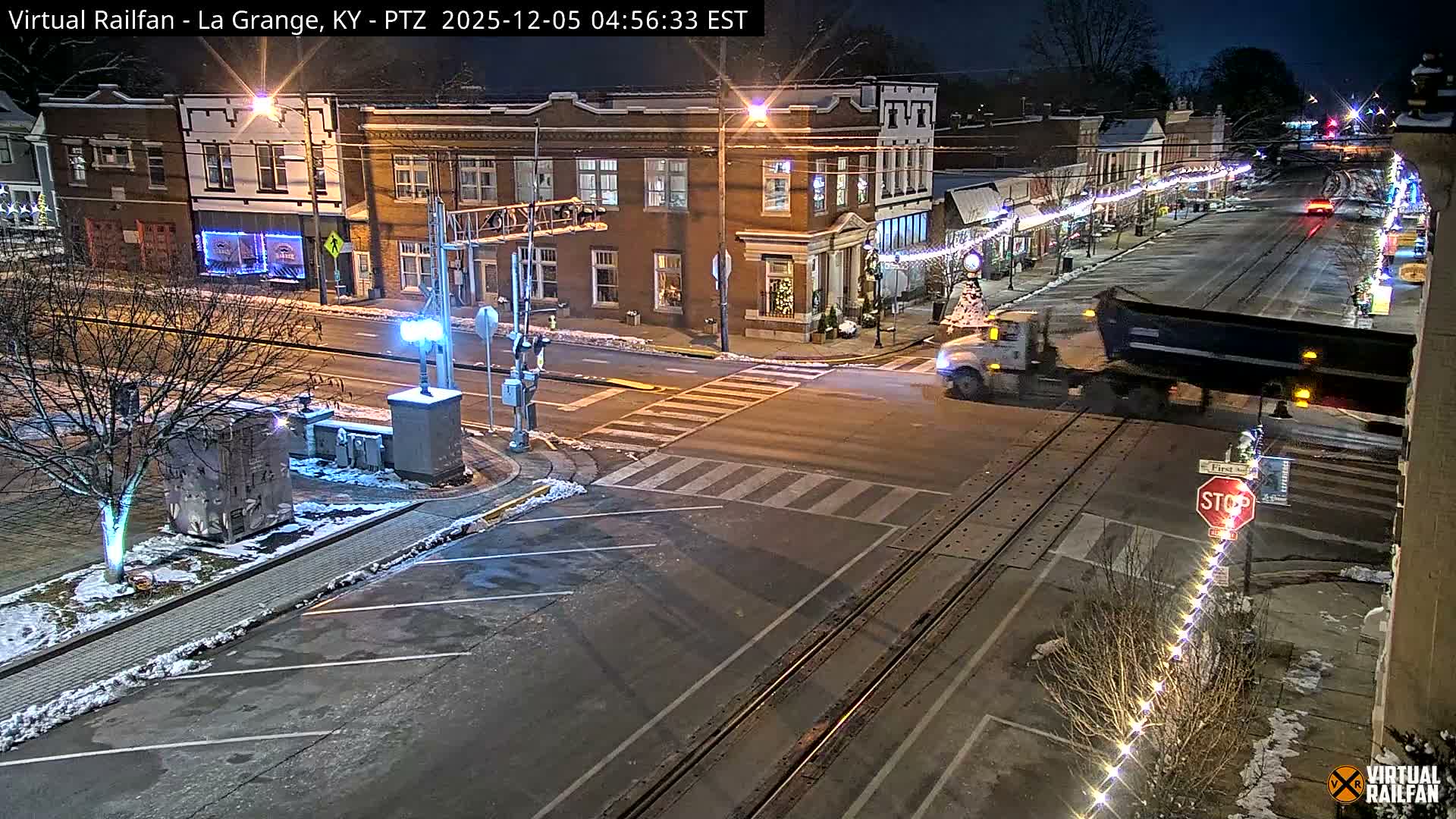 On a clear, cold winter night, a dump truck crosses snow-dusted railroad tracks on a well-lit town street adorned with decorative string lights and storefront illumination.