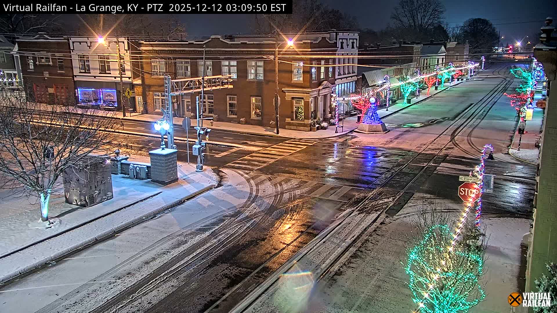 A picturesque snowy street scene at night features downtown buildings adorned with colorful holiday lights, railroad tracks traversing the wet asphalt, and trees dusted with snow under a dark sky.