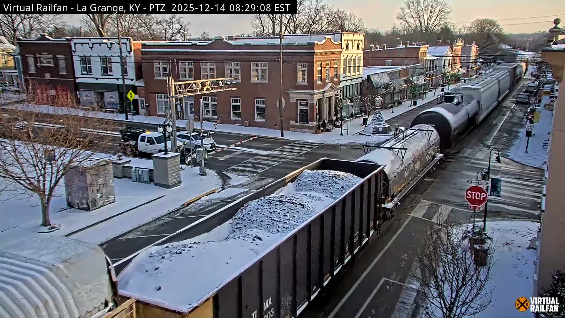 A picturesque snowy street scene at night features downtown buildings adorned with colorful holiday lights, railroad tracks traversing the wet asphalt, and trees dusted with snow under a dark sky.