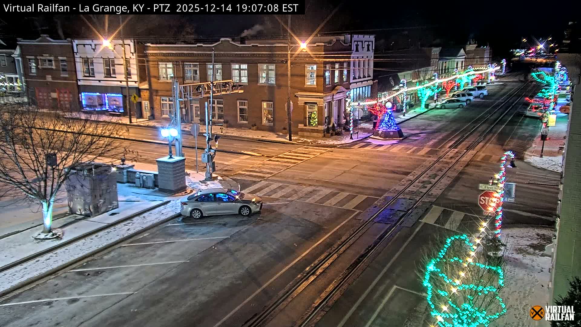 A picturesque snowy street scene at night features downtown buildings adorned with colorful holiday lights, railroad tracks traversing the wet asphalt, and trees dusted with snow under a dark sky.