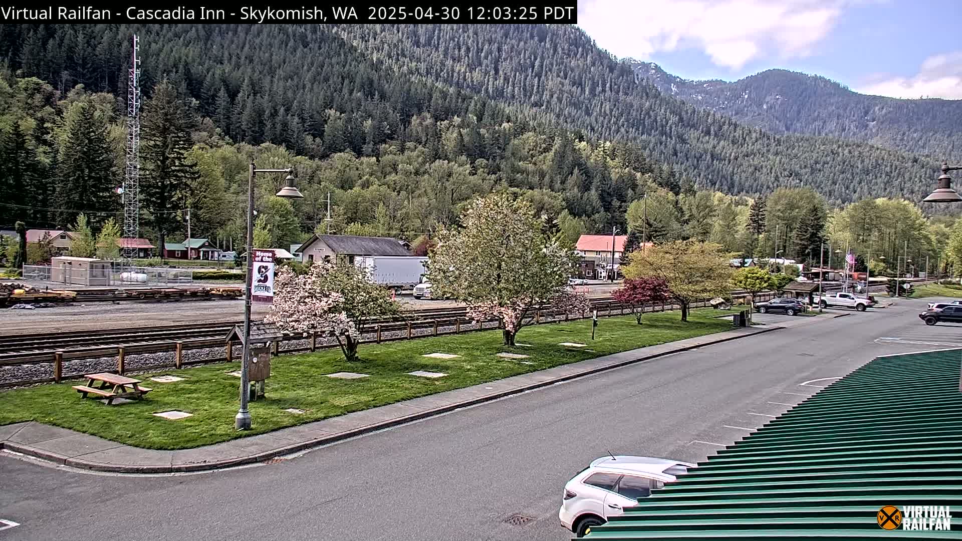 A sunny view of a small town nestled in a valley beside a mountain range, with a grassy area, trees in bloom, train tracks, and several parked cars.