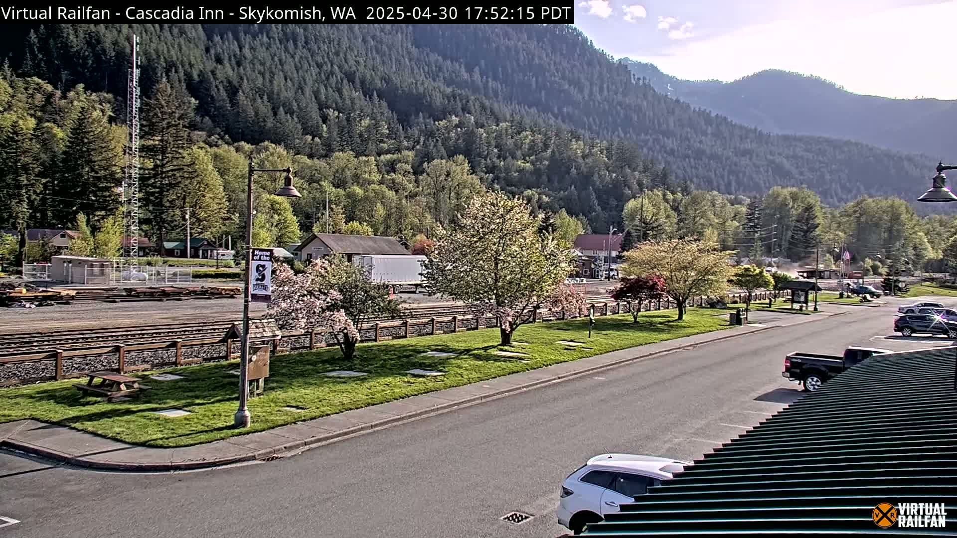 A sunny view of a town nestled in a valley beside a mountain range, featuring a railway, a road, and flowering trees.