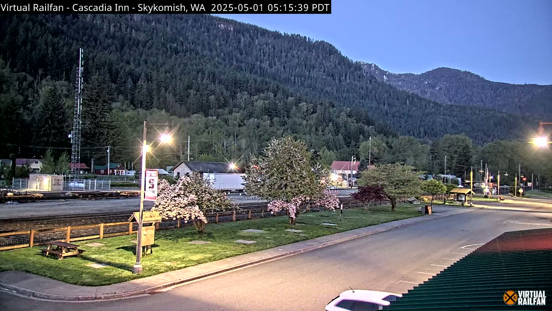 A small town nestled in a valley beneath a forested mountain range at dusk, illuminated by streetlights, features flowering trees and a picnic table near a railway line.