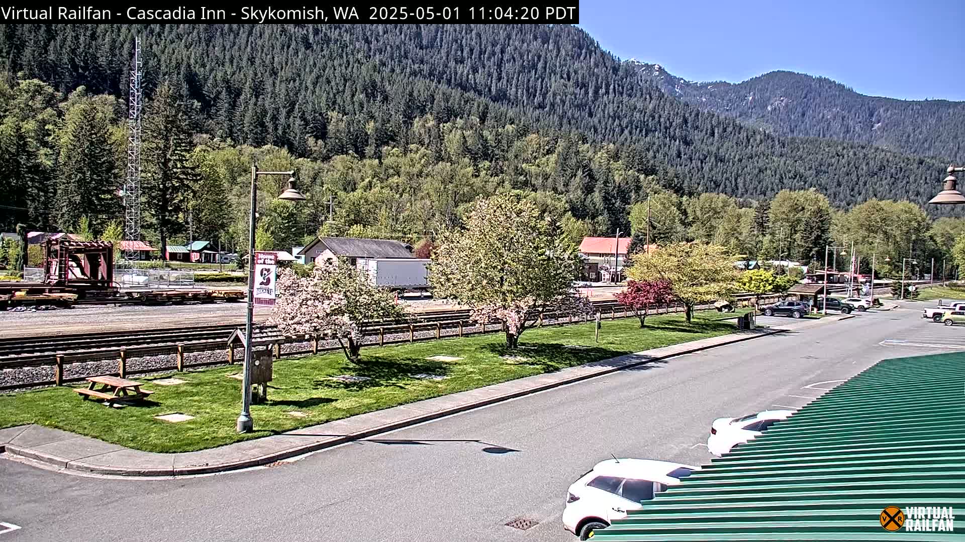 A sunny view of a town nestled in a valley, with a railway track, blossoming trees, and several parked cars visible.