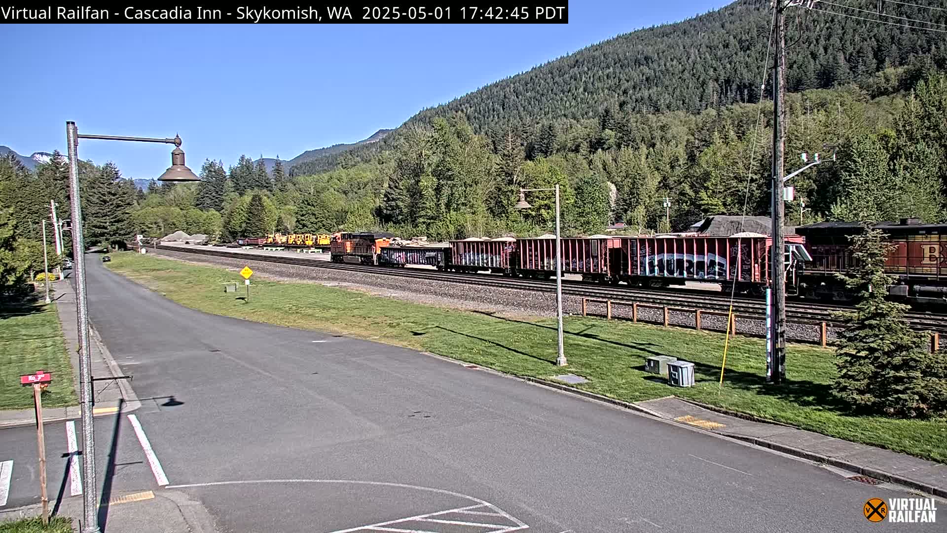 A long freight train sits on a railway track next to a road, with a background of lush green forest and mountains under a clear, sunny sky.