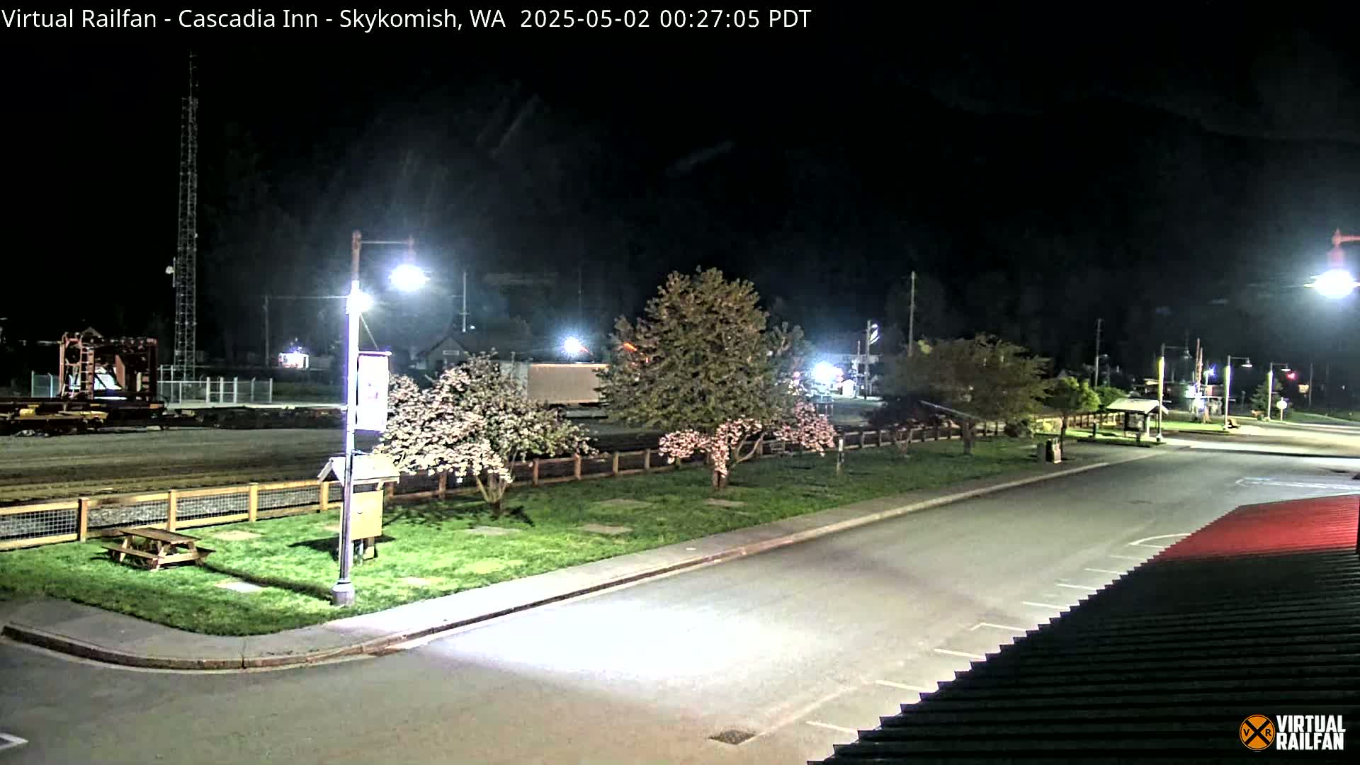 A nighttime view of a paved area with trees in blossom, a picnic table, and a fenced-off section in the background.