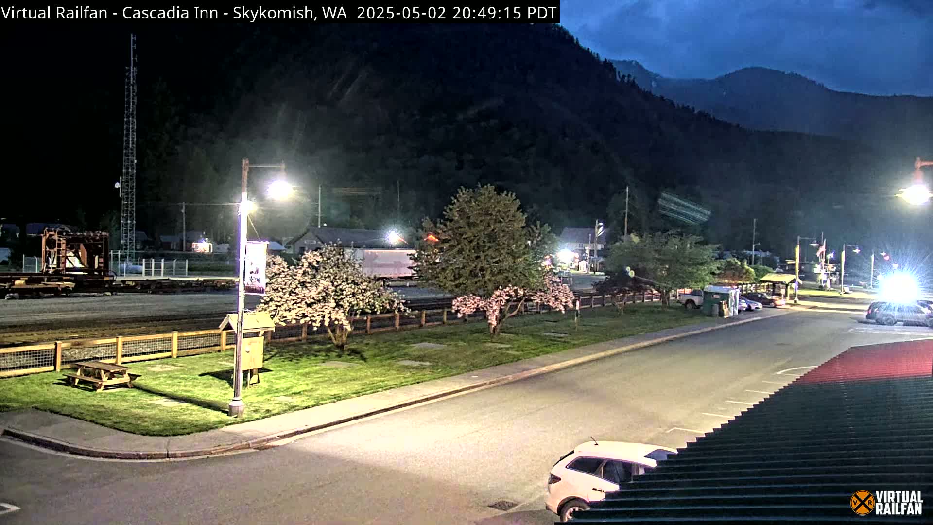 A nighttime view of a small town near a mountain, featuring a parking lot with a few cars, some flowering trees, and a picnic table in a grassy area.