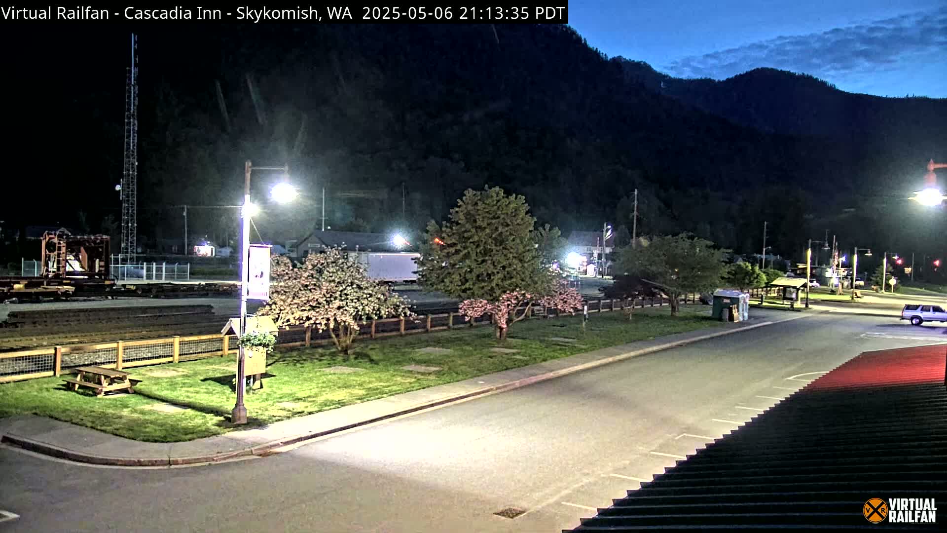 A nighttime view of a parking lot and grassy area with flowering trees, bordered by a roadway, a rail yard, and a mountainside under a twilight sky.