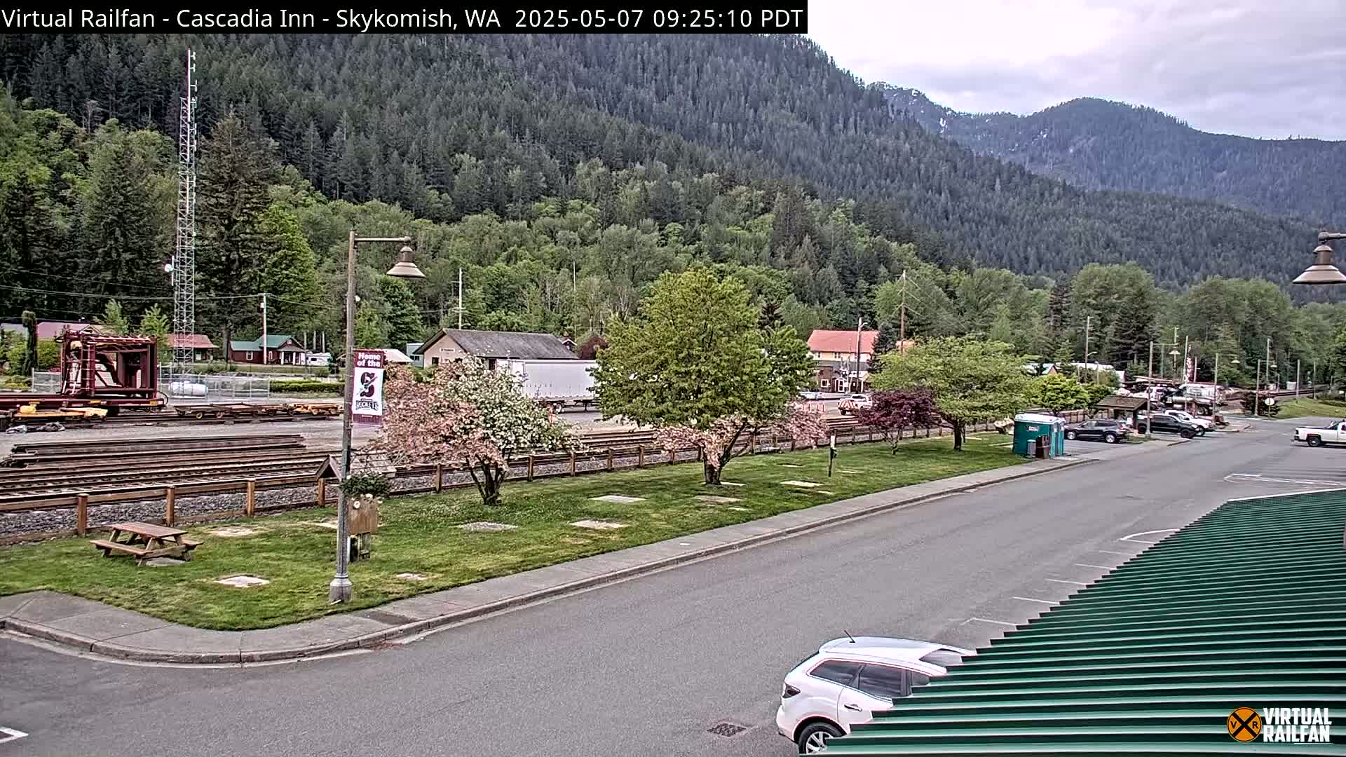 A partly cloudy day reveals a small town nestled in a valley surrounded by lush green mountains, with railroad tracks and a few vehicles visible near a grassy area.