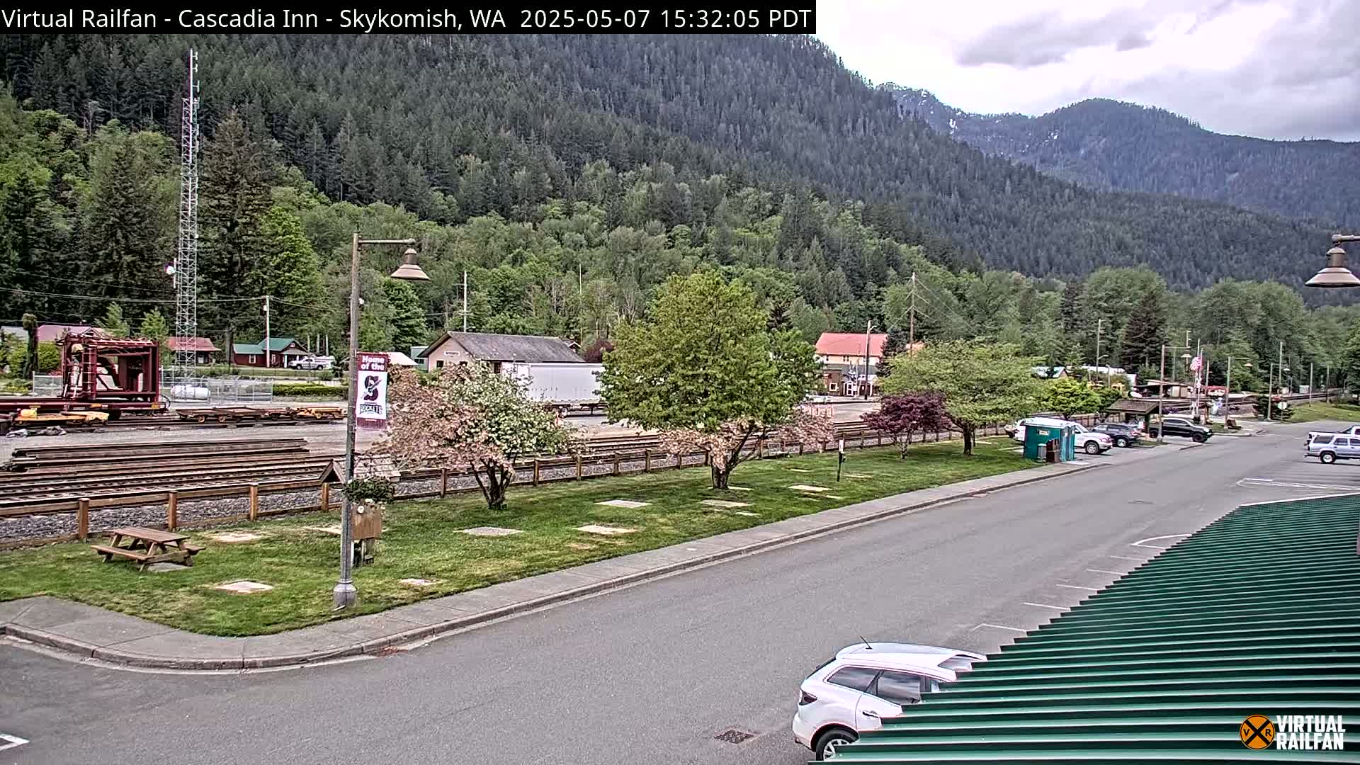 A partly cloudy day reveals a view of railroad tracks, a parking lot, and several vehicles near a small town nestled in a valley surrounded by lush green mountains.