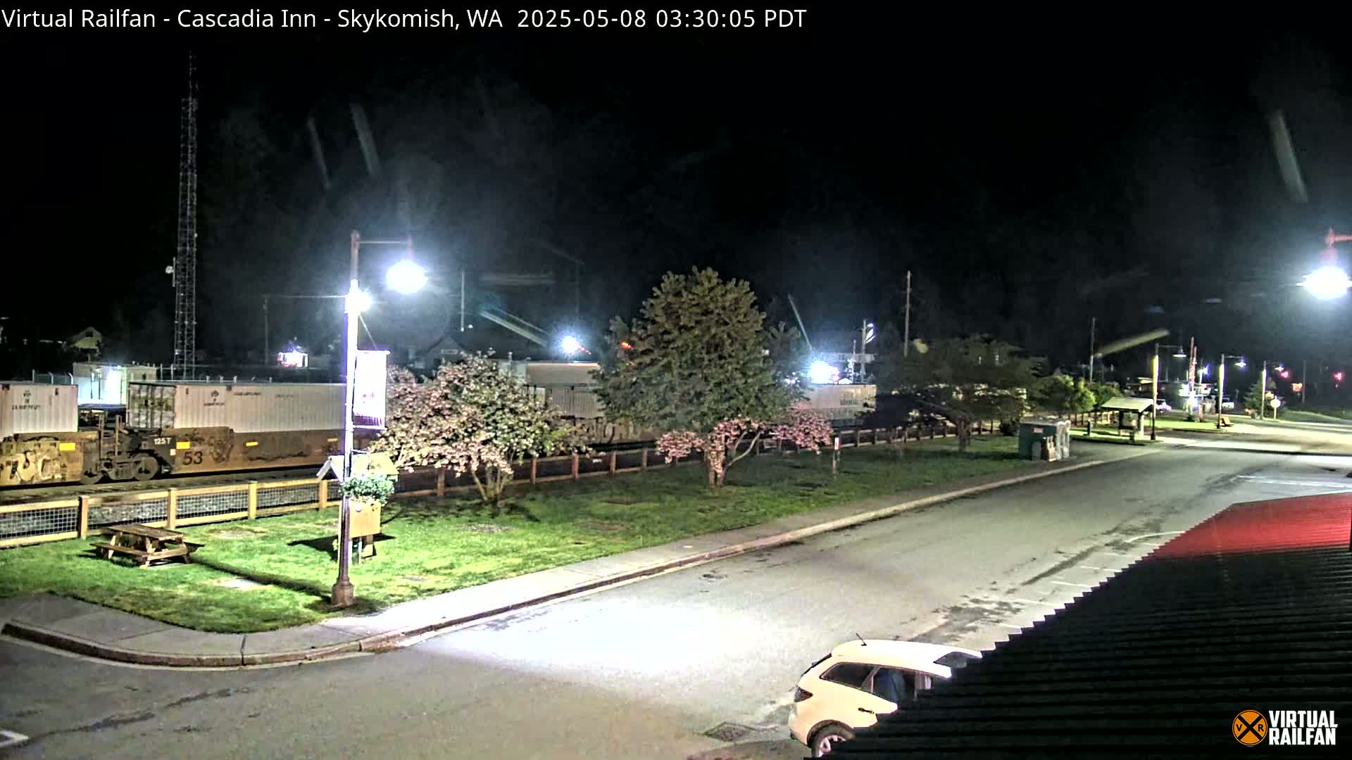 At night, a train is parked next to a grassy area with flowering trees and streetlights, and a car is visible in a parking lot nearby.