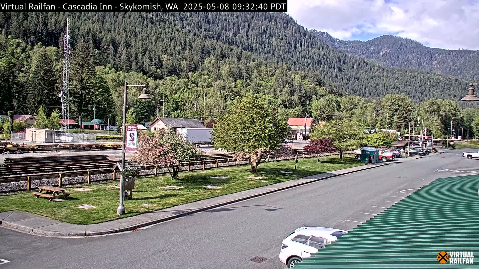 A partly cloudy day reveals a mountainside town with train tracks, parked cars, and flowering trees alongside a road.