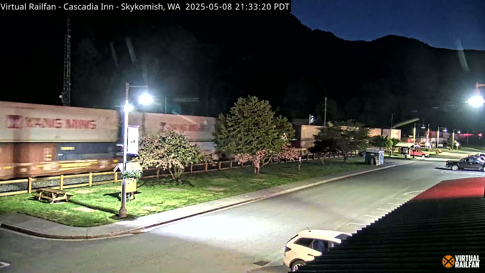 A long freight train passes by a parking lot at night under dark, clear skies, with several cars parked and a few flowering trees visible.