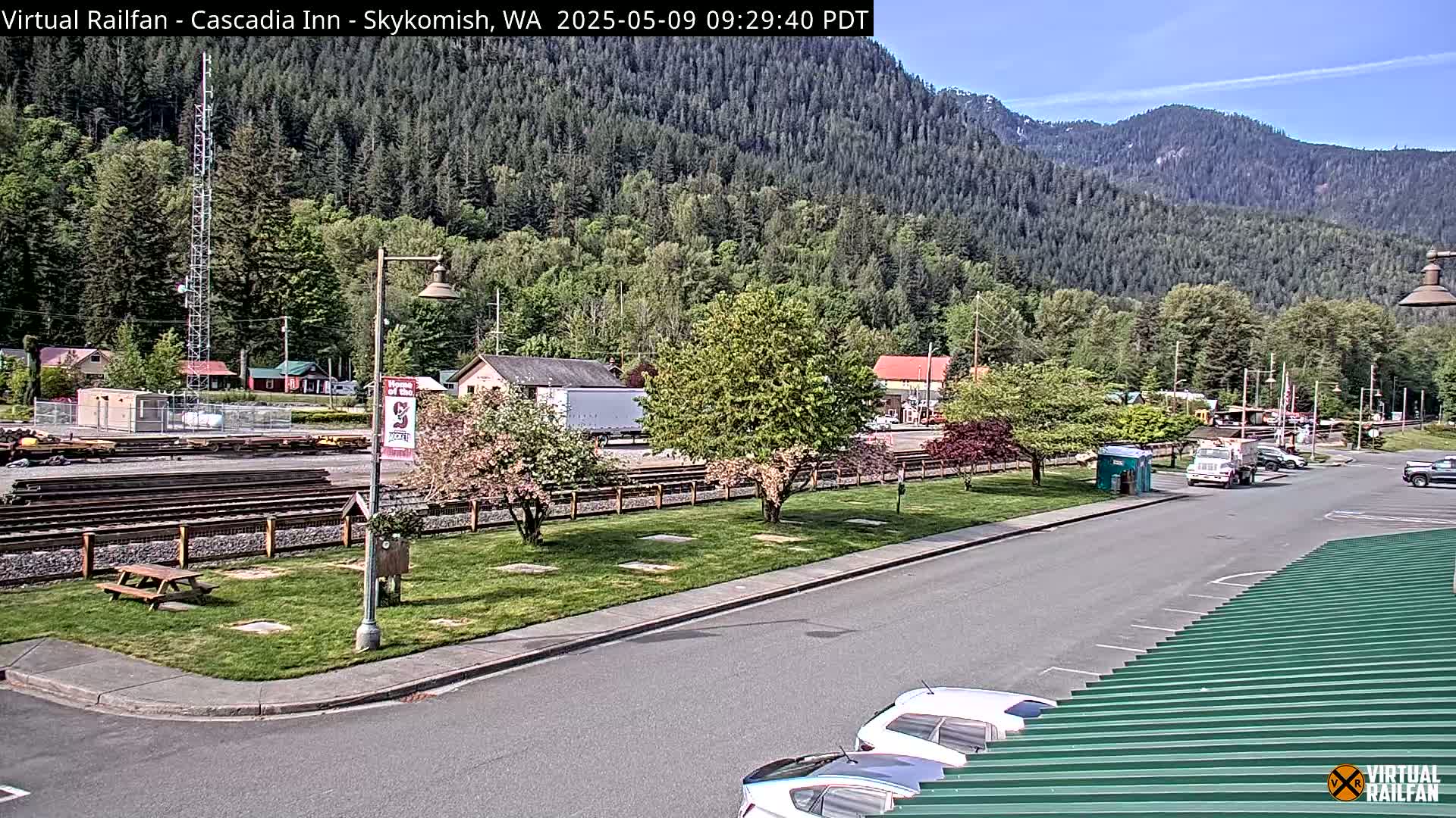 A sunny view of a parking lot and train tracks, bordered by green grass, trees, and buildings, with a mountain range in the background.
